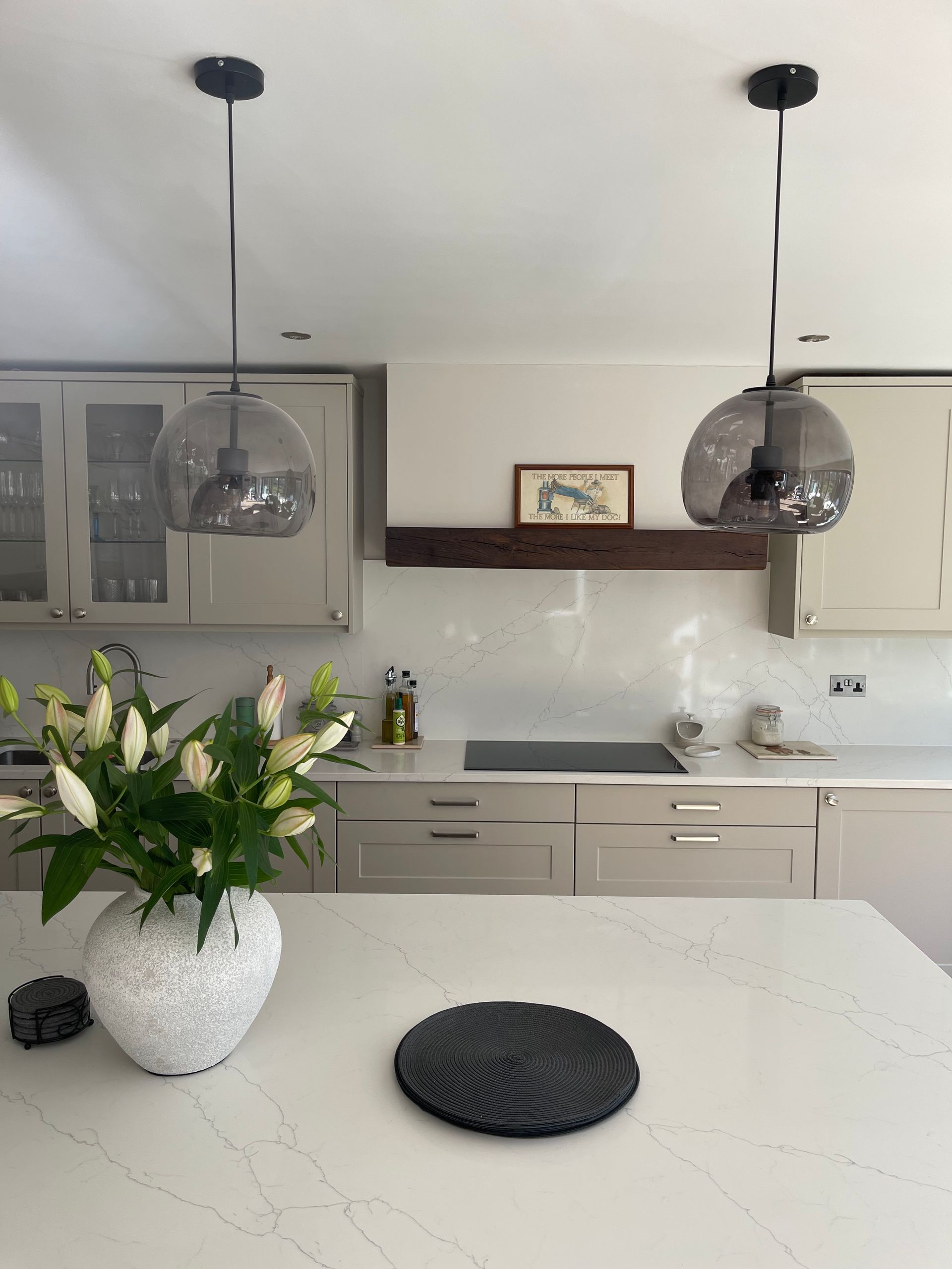 A kitchen island with a white vase of lilies and a dark coaster, beneath two grey glass pendant lights and modern cabinets.