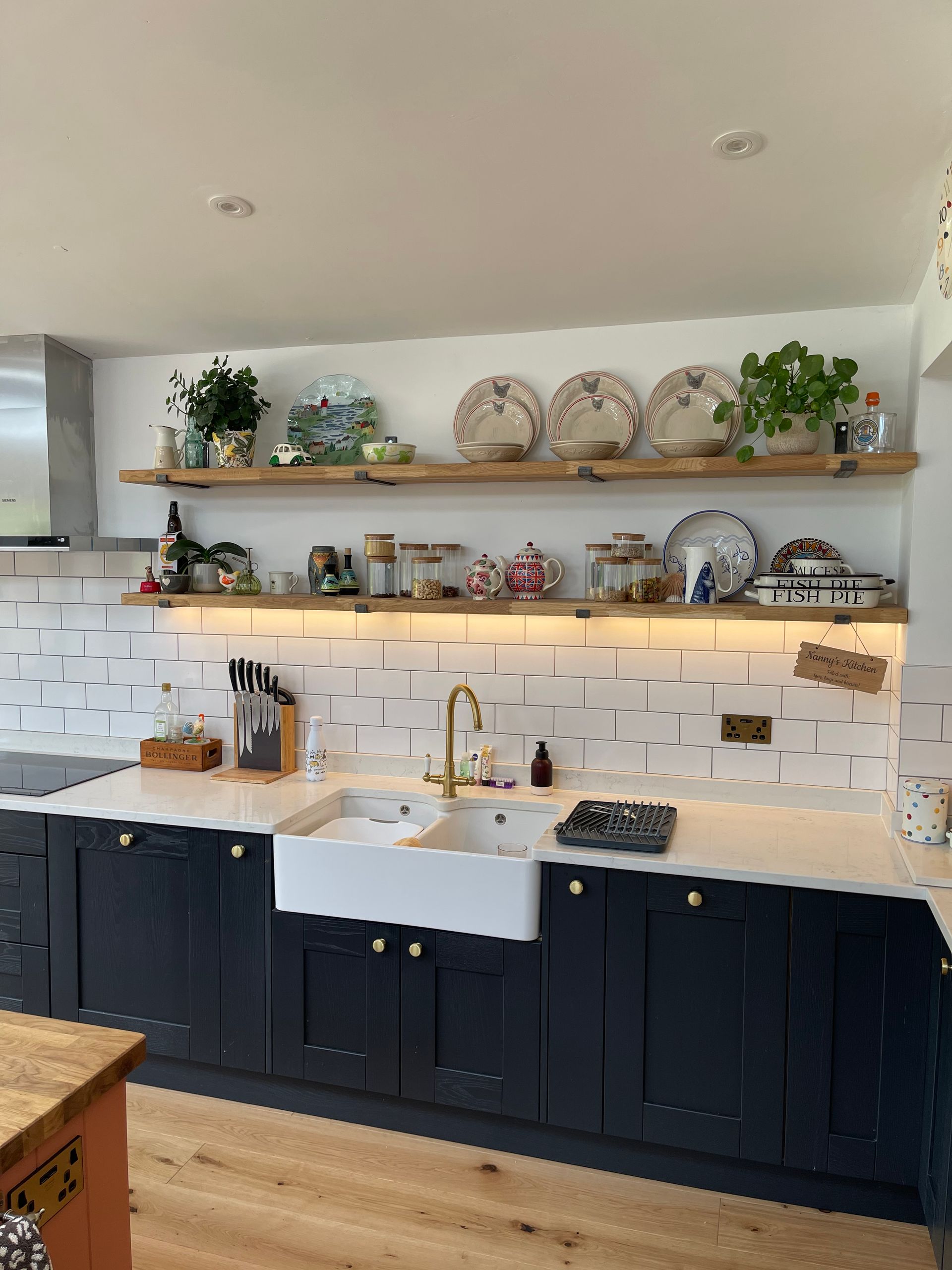 A kitchen featuring dark blue cabinets, white countertops, a farmhouse sink, and wooden shelves filled with dishware.