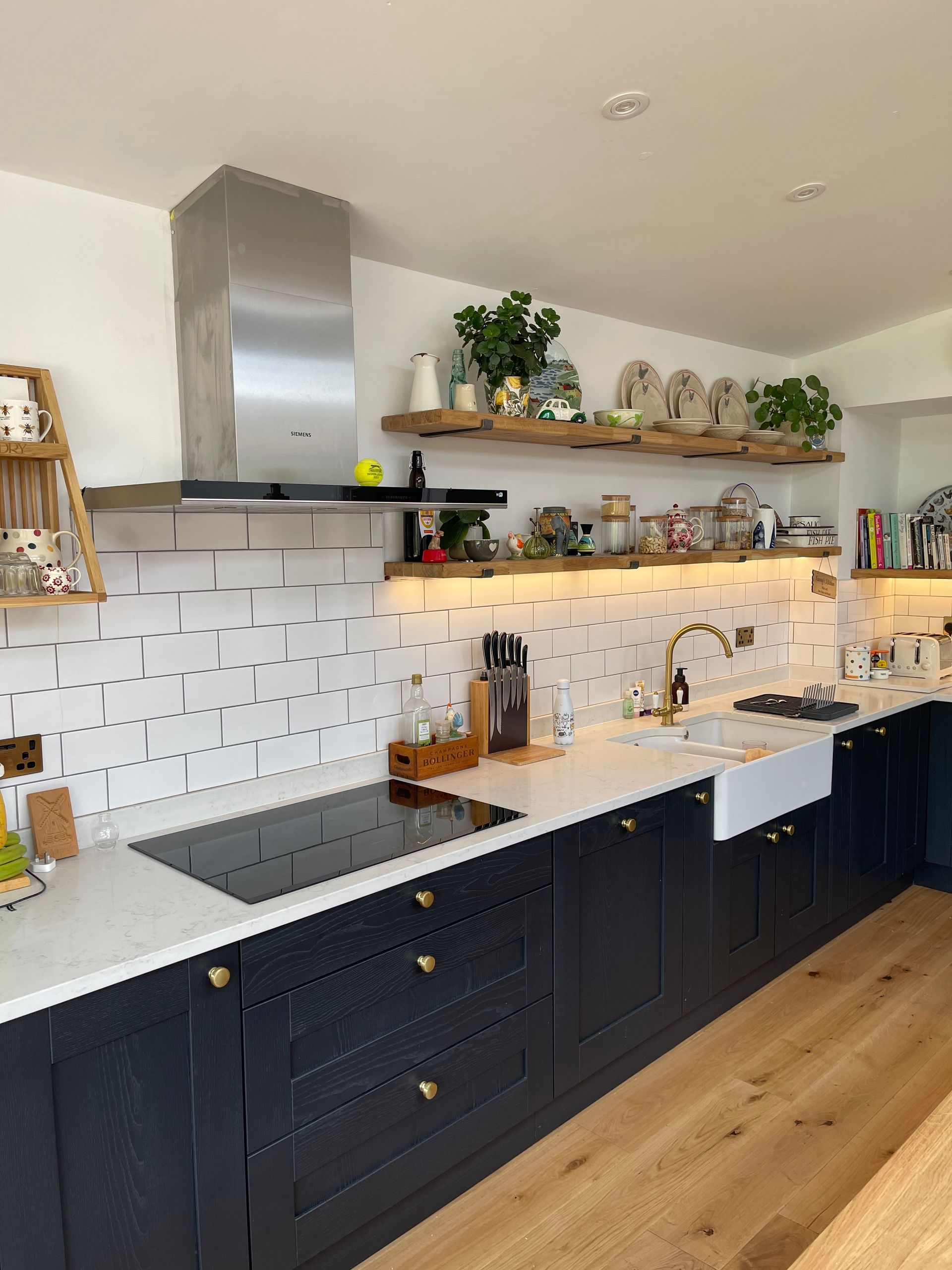 A modern kitchen with dark blue cabinets, white countertops, subway tile, and open wood shelving under warm lights.