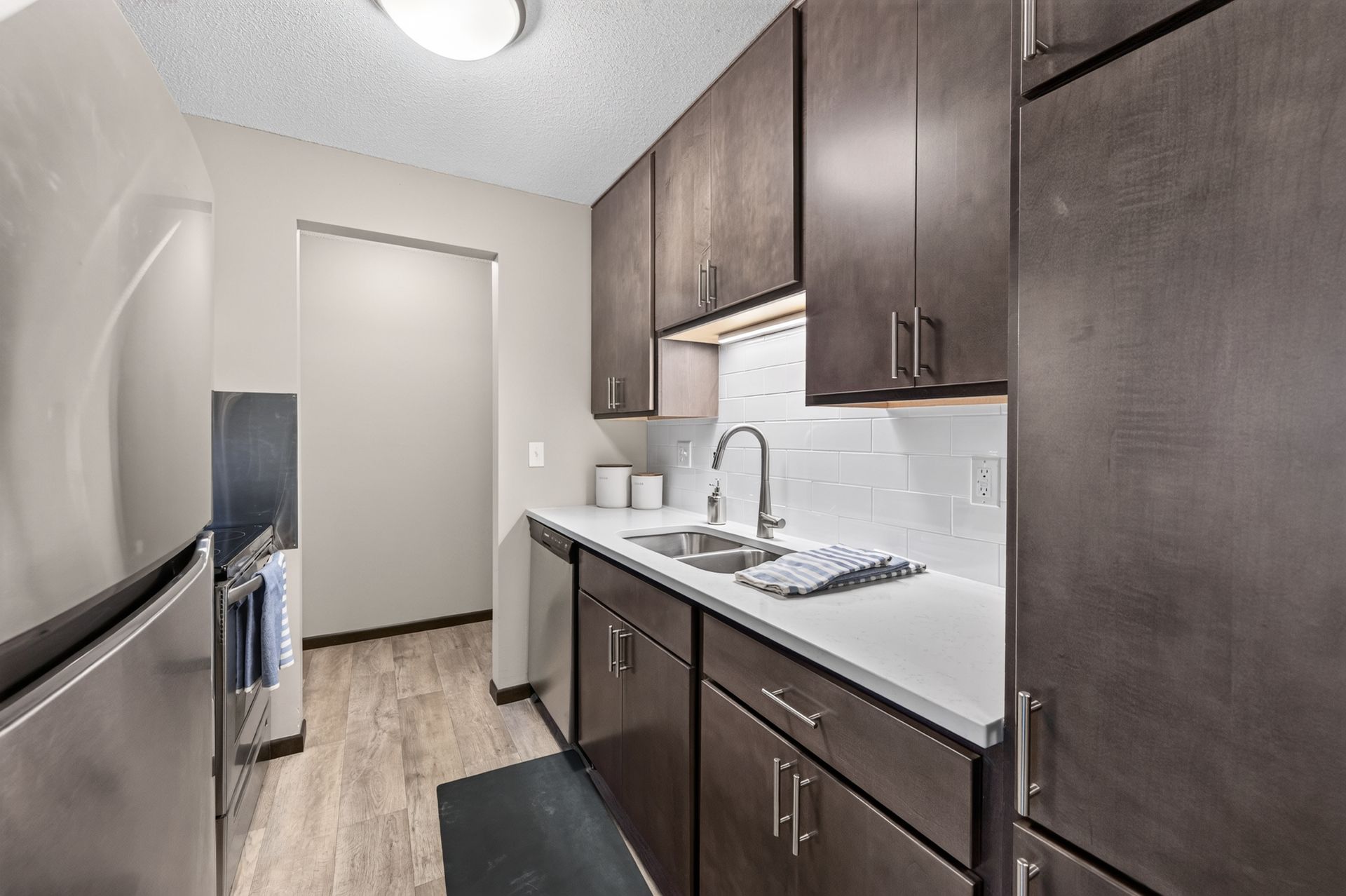 Narrow kitchen with dark wood cabinets, white countertops, stainless sink, and light wood flooring