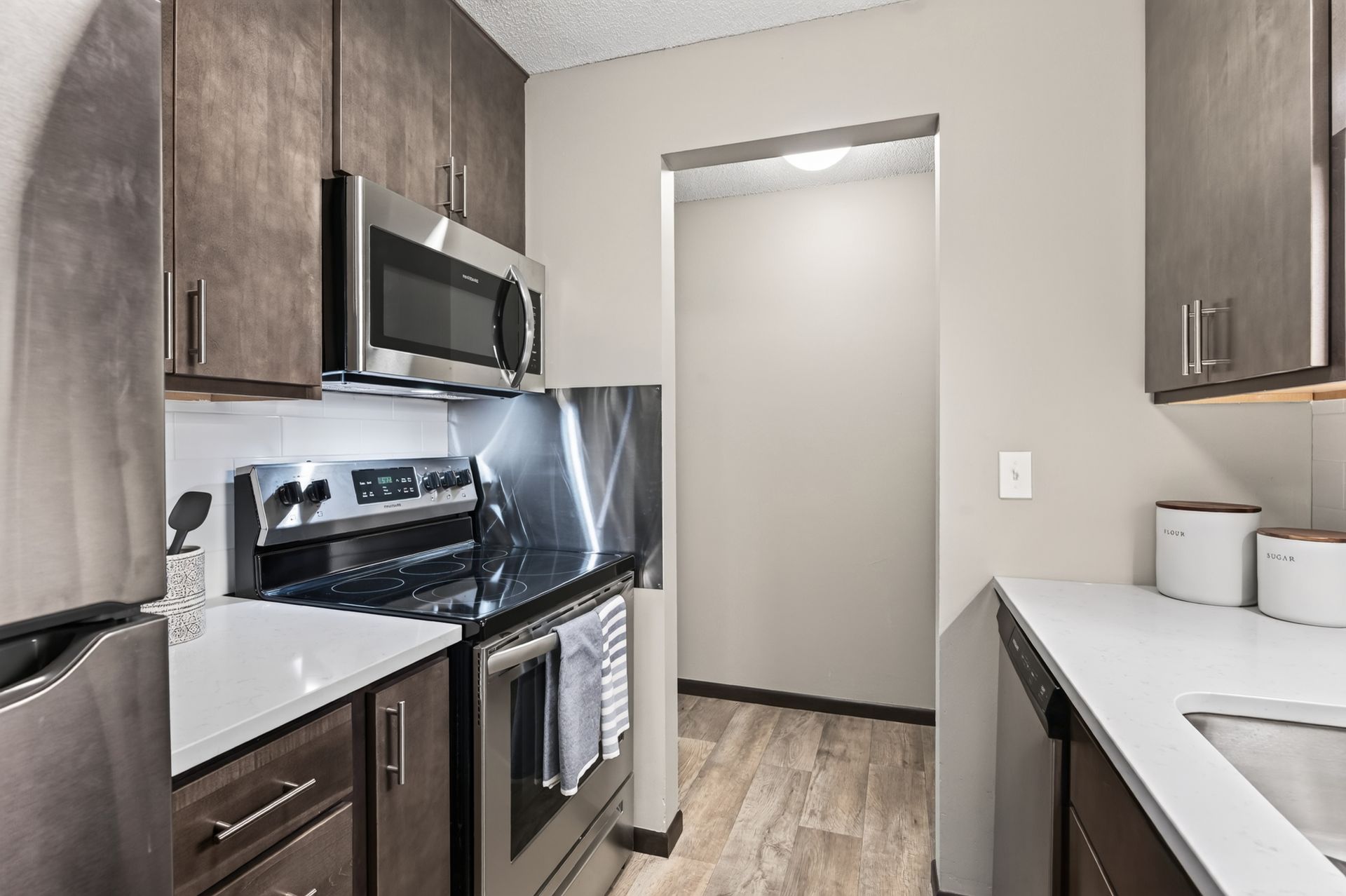 Narrow modern kitchen with dark cabinets, stainless appliances, and light countertops.