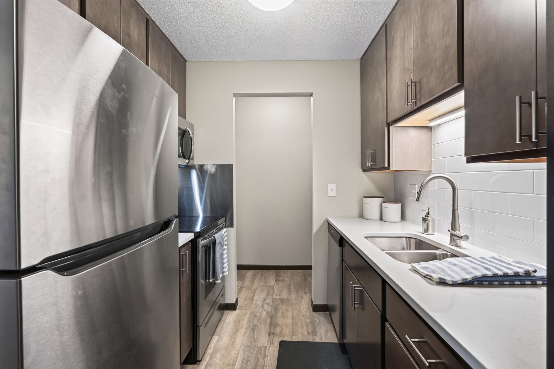 Narrow galley kitchen with stainless appliances, dark cabinets, white counters, and wood-look flooring
