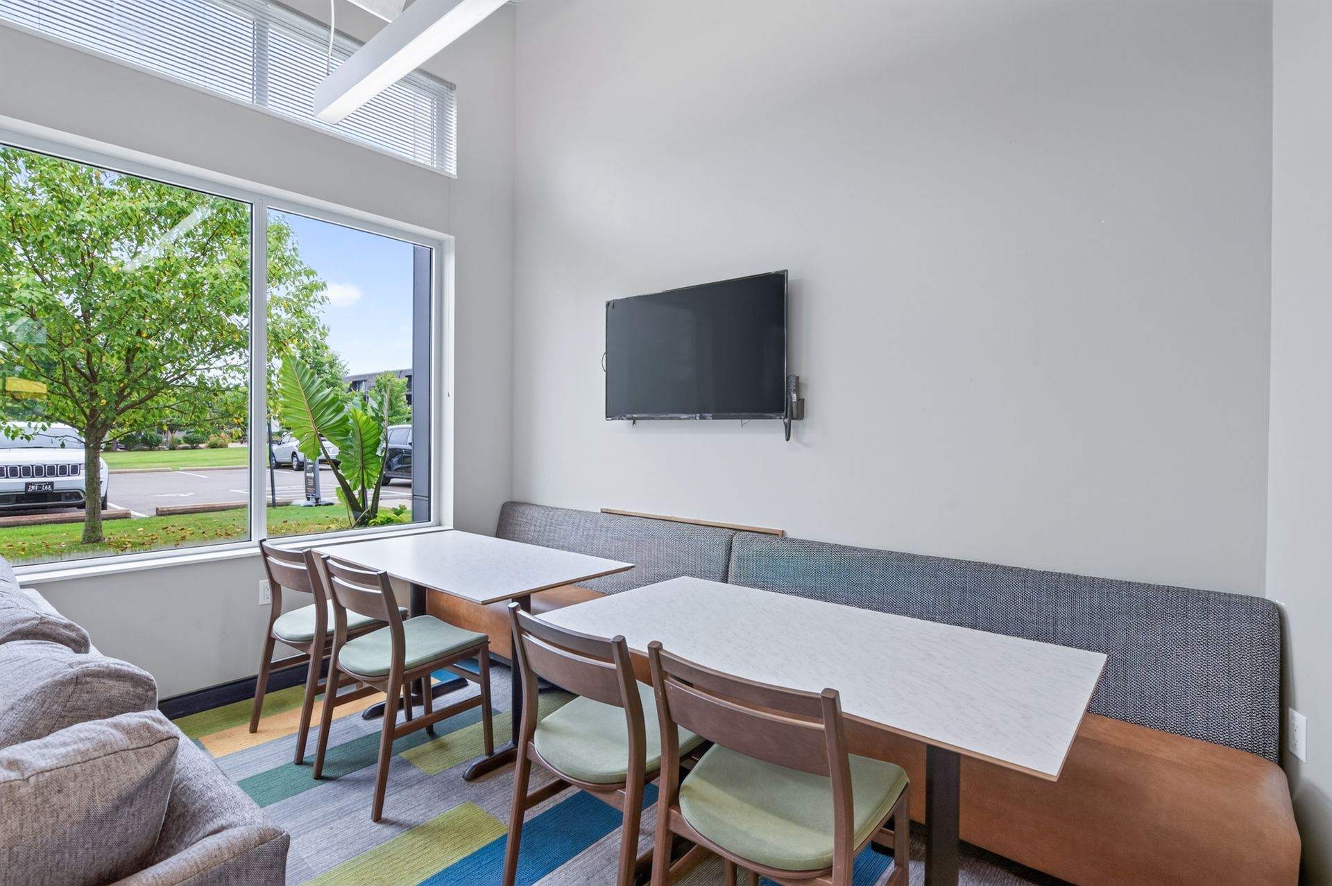 Modern meeting room with long white tables, wooden chairs, wall-mounted TV, and large windows overlooking trees