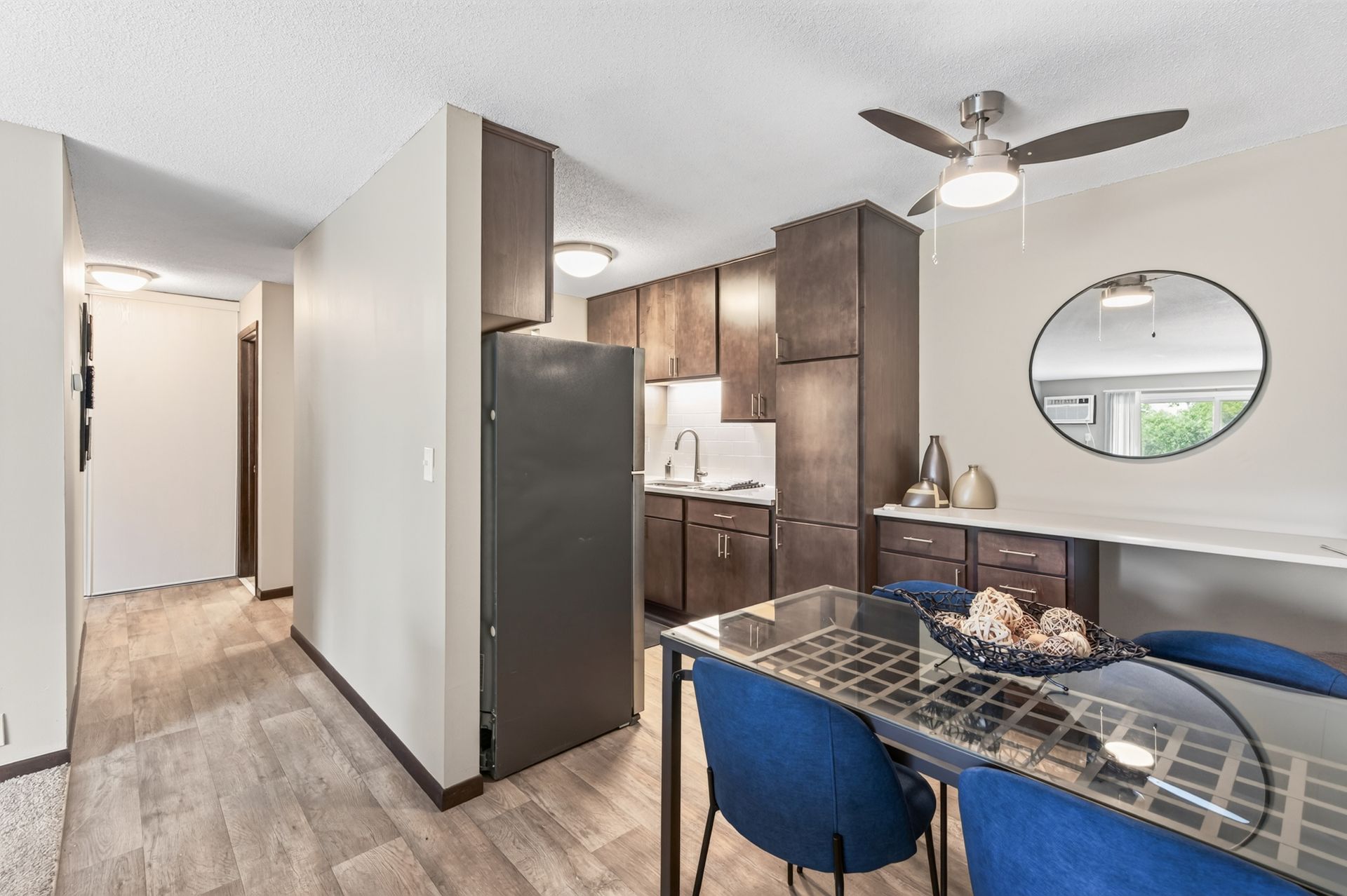Modern apartment kitchen and dining area with dark cabinets, stainless fridge, blue chairs, and hallway view