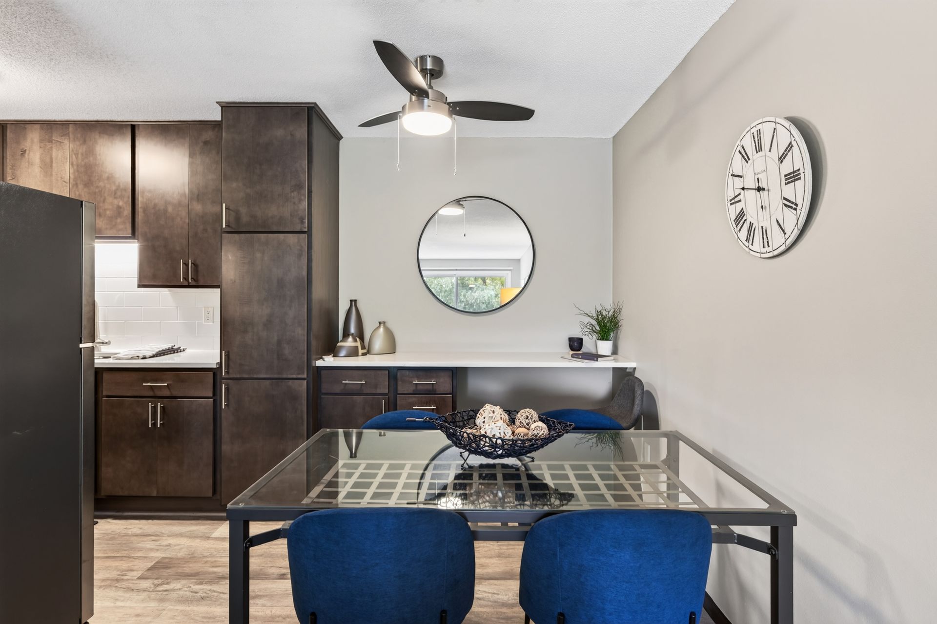 Modern dining area with glass table and blue chairs beside a kitchen, mirror, and wall clock