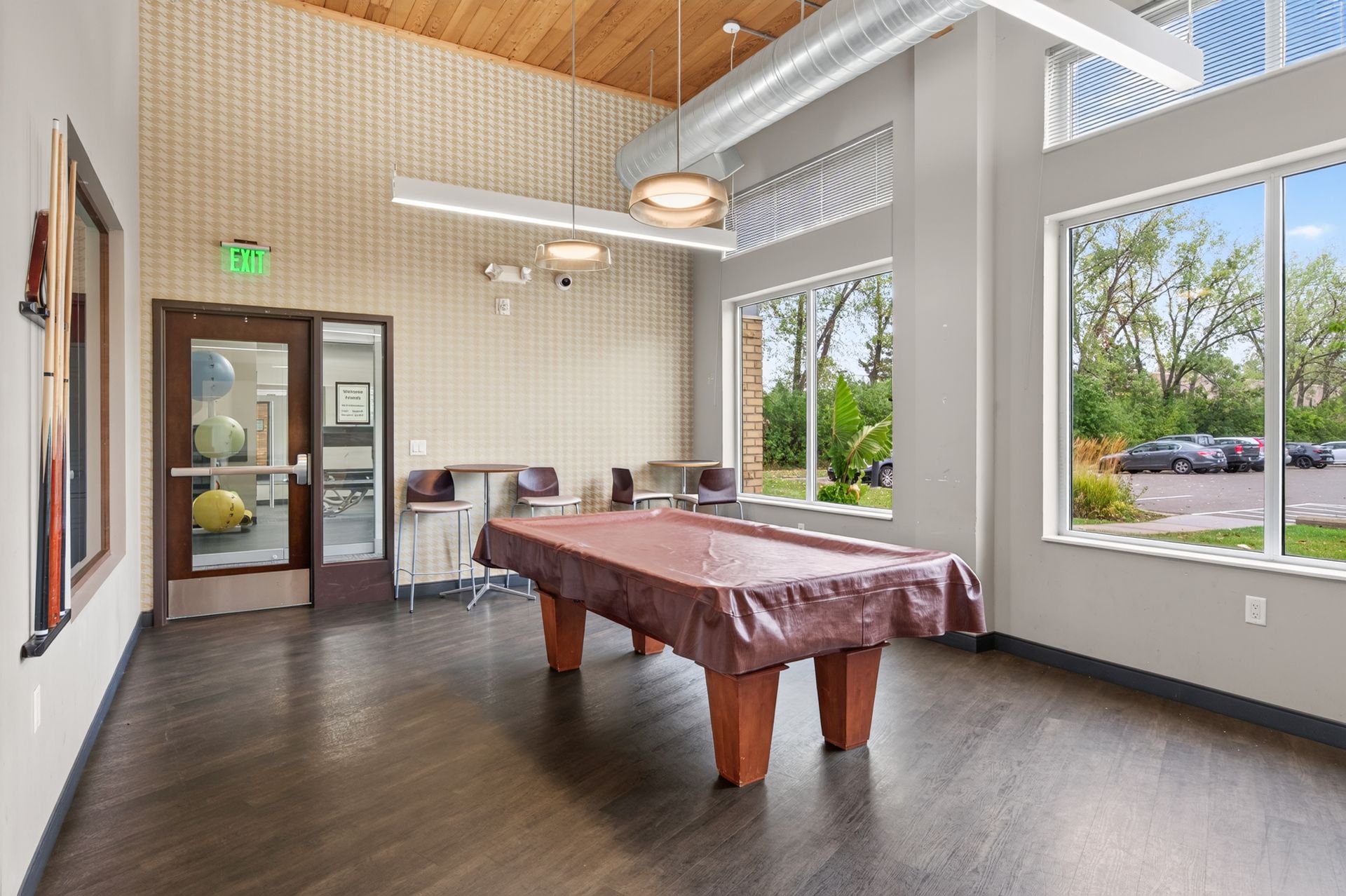 Bright community room with pool table, large windows, wood ceiling, and chairs by the door