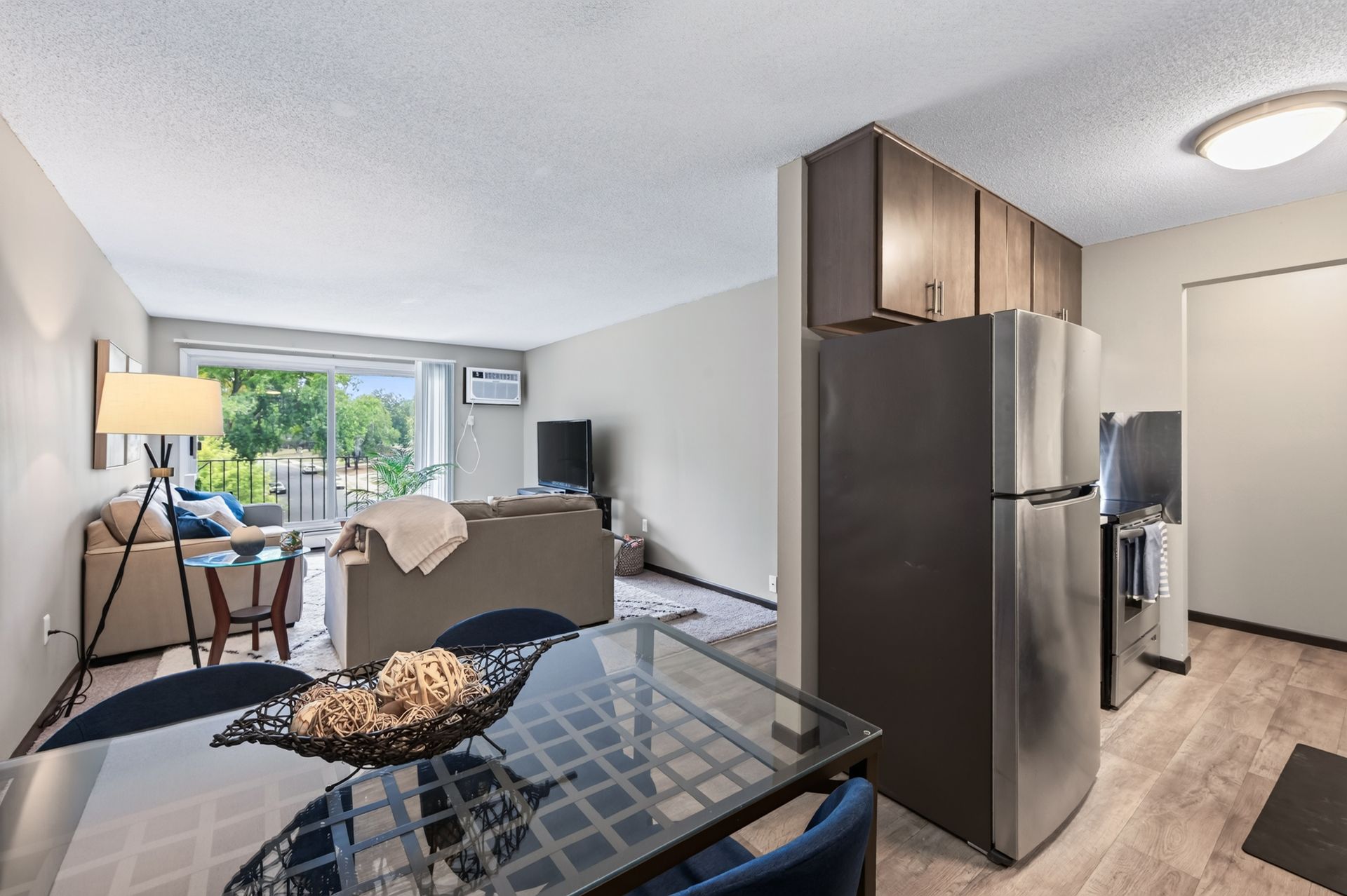 Modern apartment living room and kitchen with glass dining table, sofa, and stainless steel refrigerator.