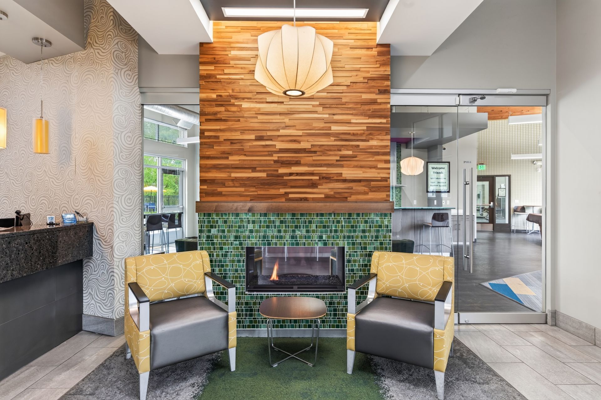 Modern lobby seating area with two gray chairs, a fireplace, wood accent wall, and green tile backdrop