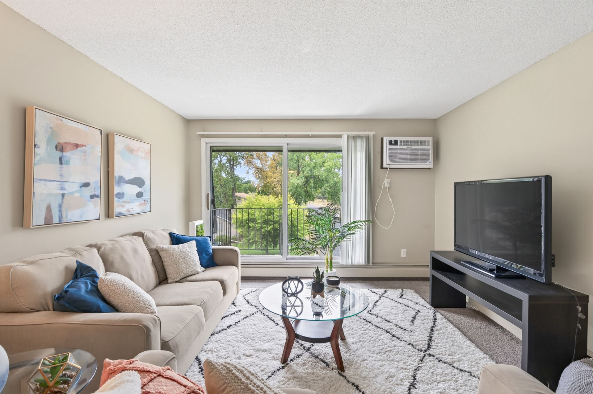 Bright living room with beige sofa, TV, coffee table, and sliding glass doors to greenery