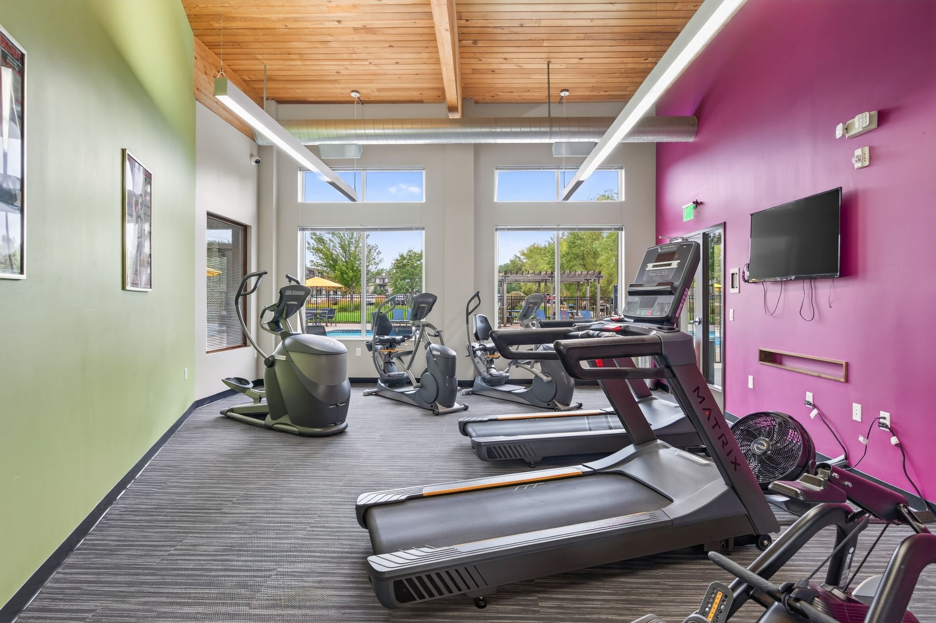 Bright gym with treadmills, exercise bikes, wall-mounted TV, and large front windows.