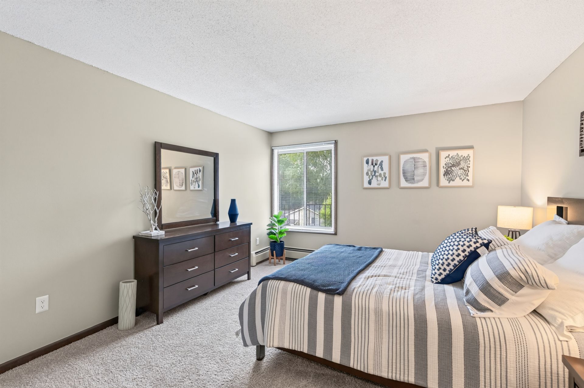 Bright bedroom with striped bed, dark dresser, wall art, and window with greenery view.