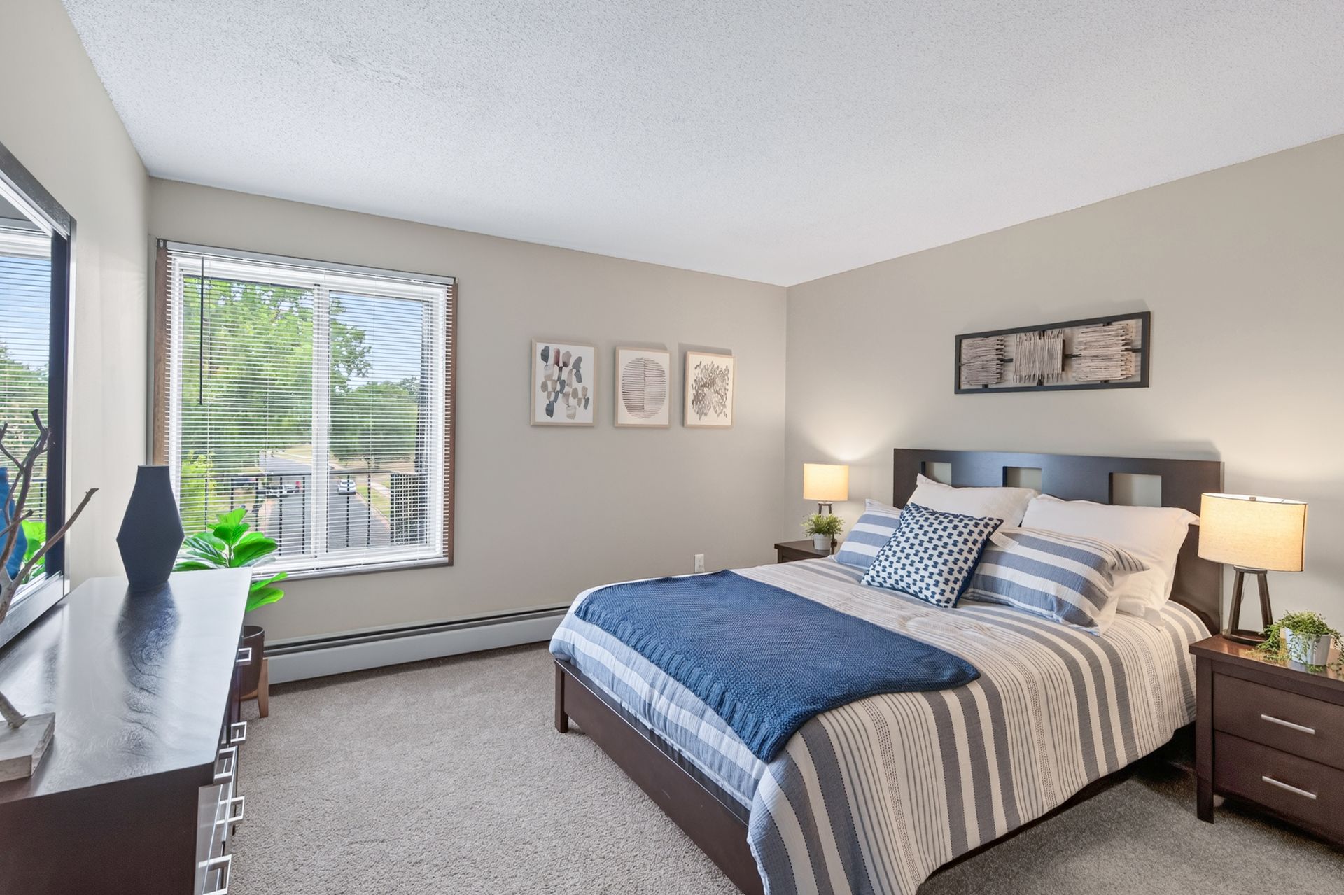 Bright bedroom with a bed, nightstand lamp, wall art, and large window with blinds