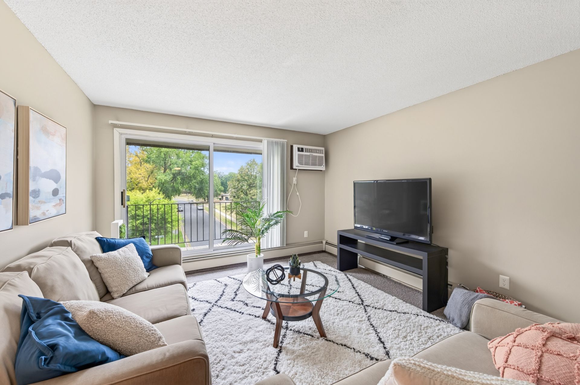 Bright living room with beige sofas, TV stand, coffee table, and sliding glass doors to a patio and greenery
