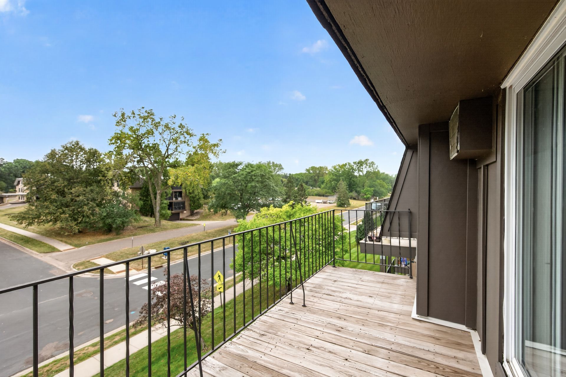 Balcony with black railing overlooking a quiet street and green trees under a blue sky