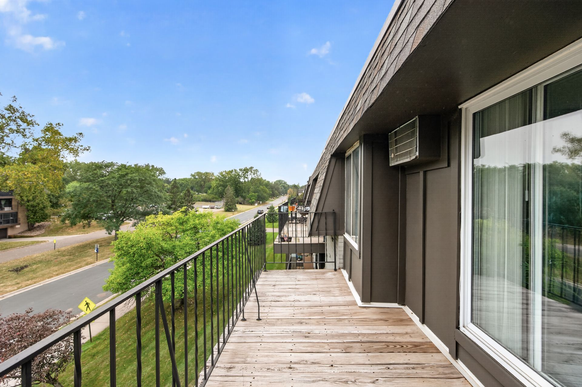 Wooden balcony walkway beside a modern building, overlooking a road and green trees under a blue sky