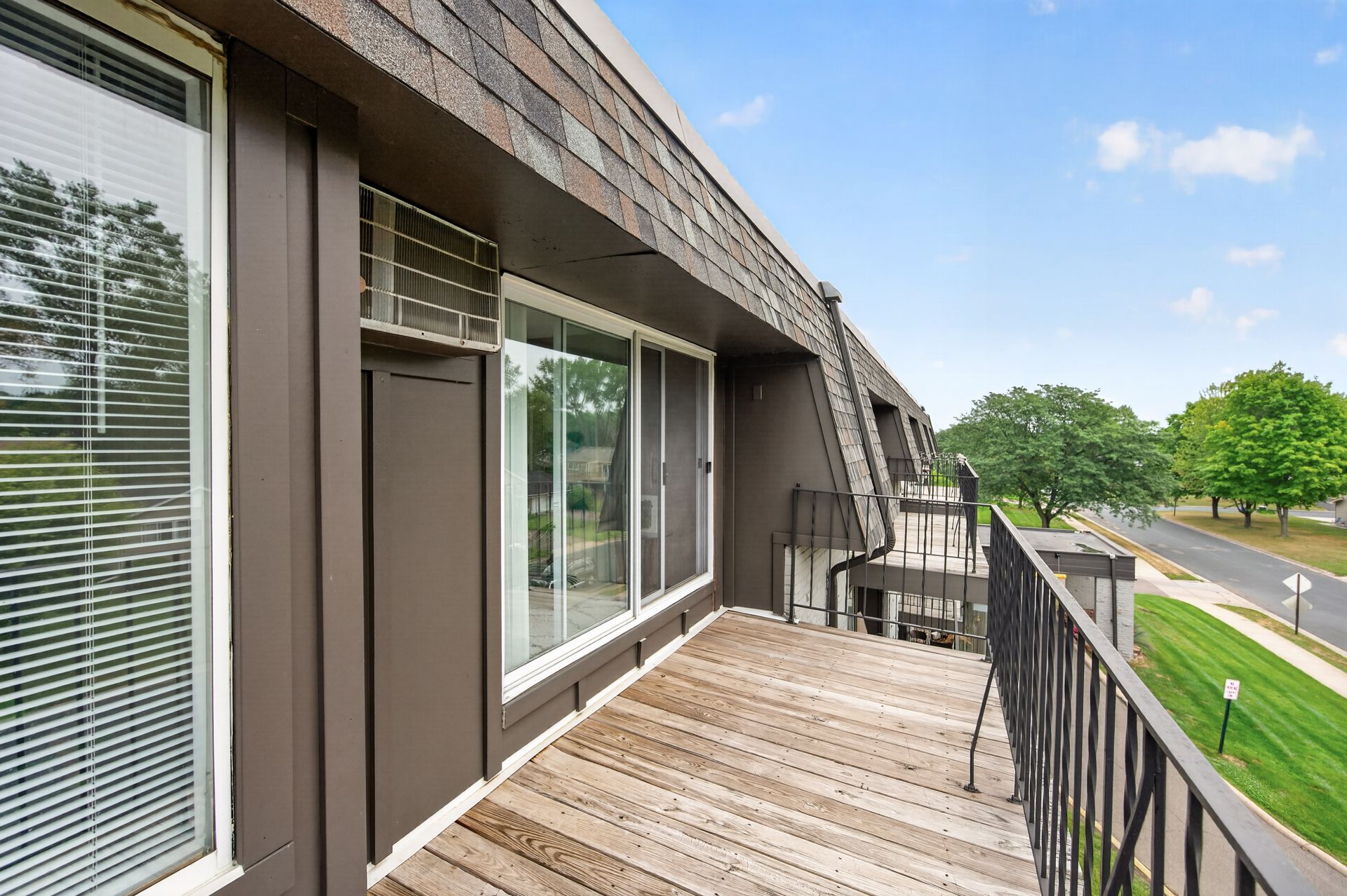 Apartment balcony with wooden deck, black railing, and sliding glass doors overlooking a road and trees