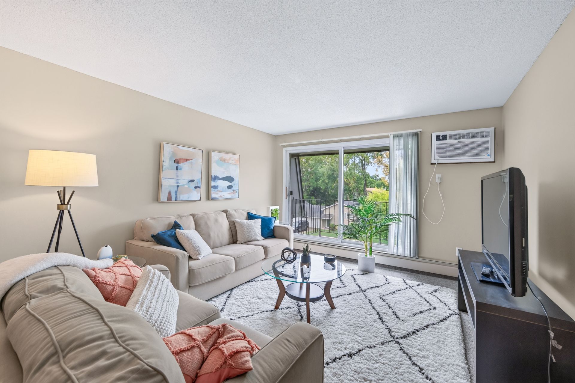 Bright living room with beige sofas, coffee table, TV, lamp, and sliding glass door to a balcony