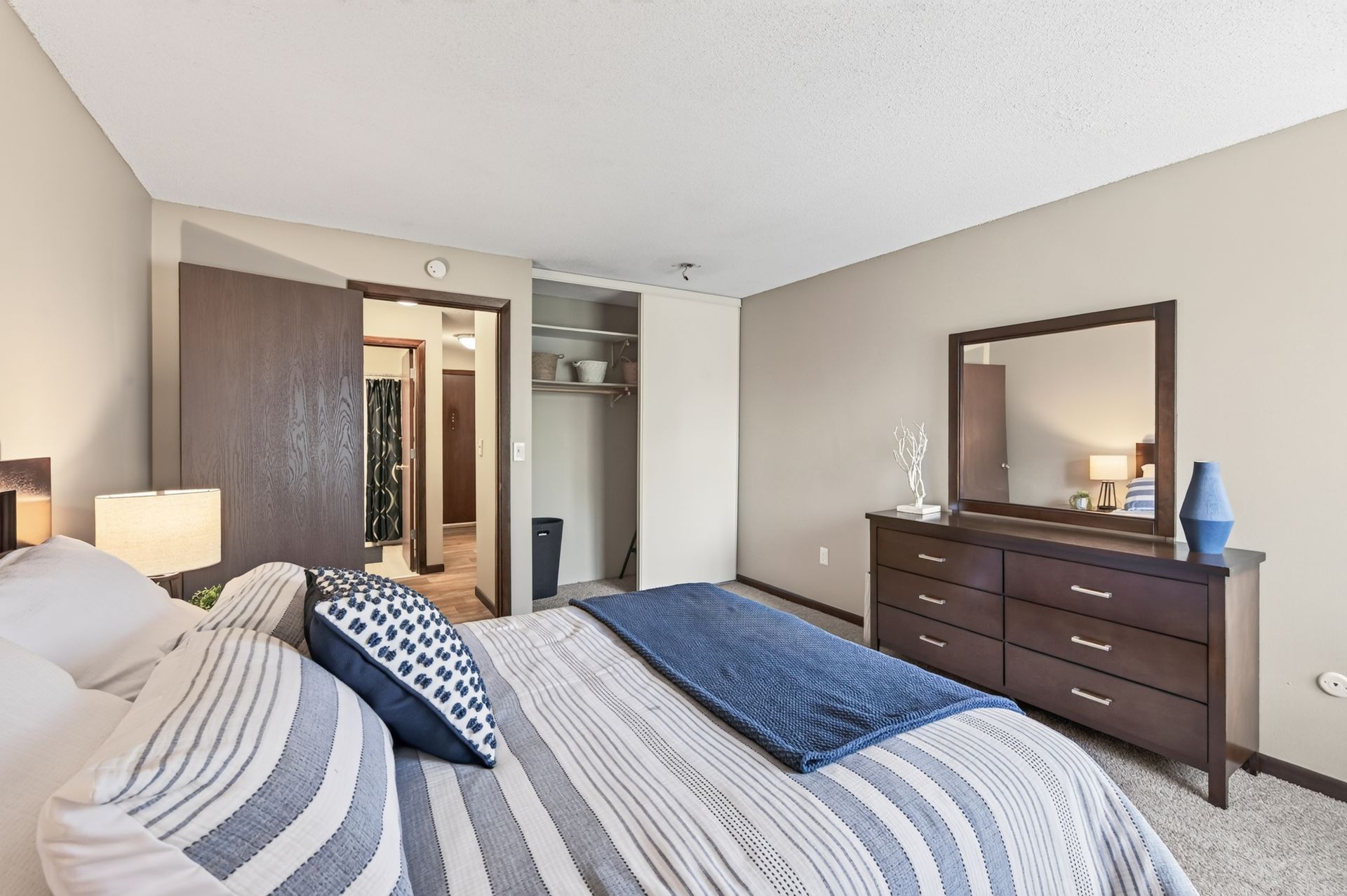Bedroom with striped bedspread, dark wood dresser with mirror, and open closet doors in a neutral-toned room