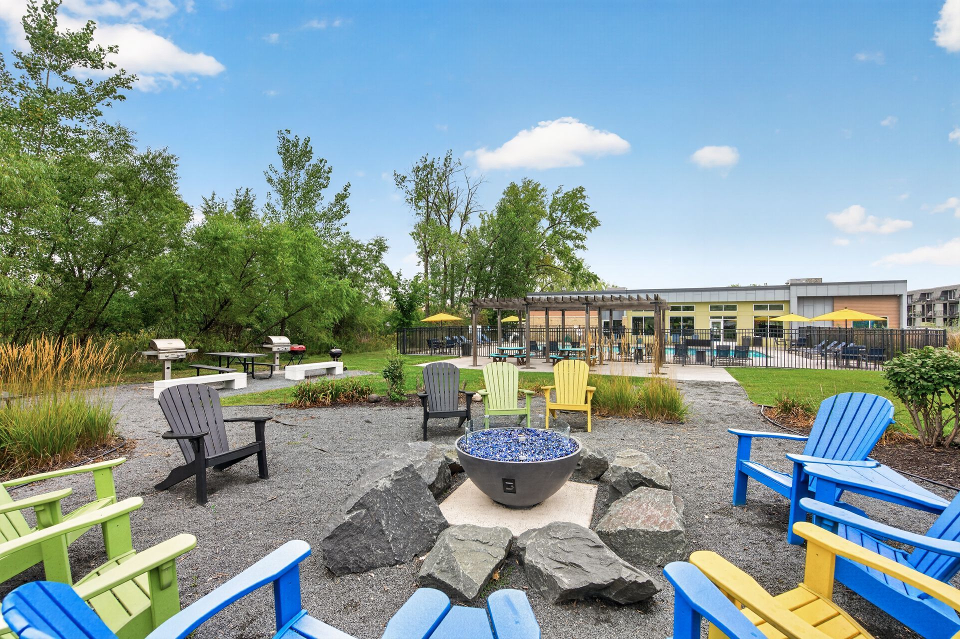 Outdoor seating area with colorful chairs around a fire pit in a landscaped park under a blue sky