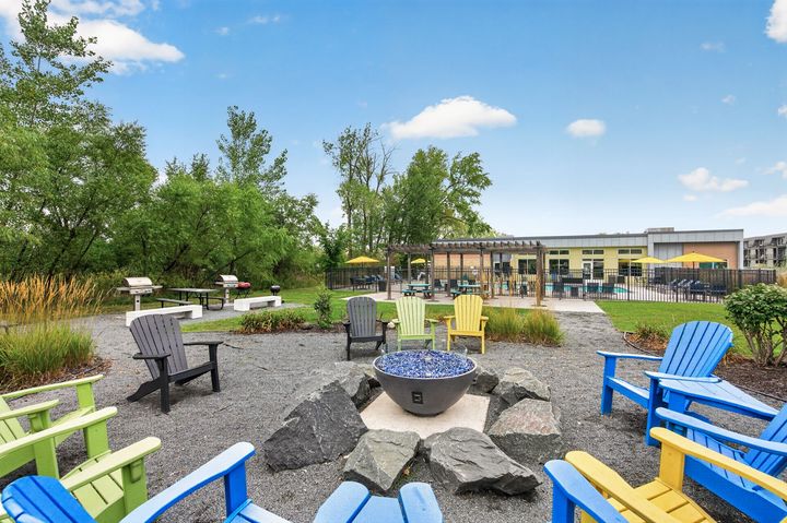 Outdoor patio with blue chairs around a stone fire pit beside a park and building.