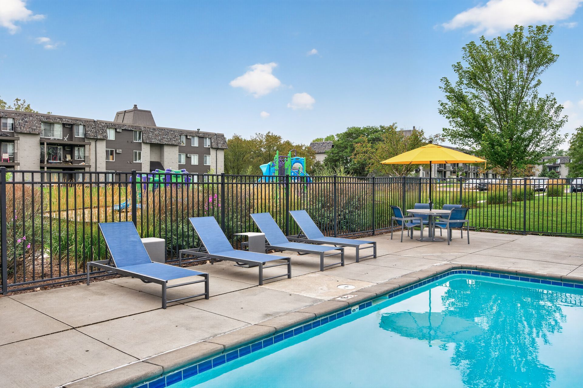 Poolside patio with blue lounge chairs, tables, yellow umbrella, and apartment buildings in the background