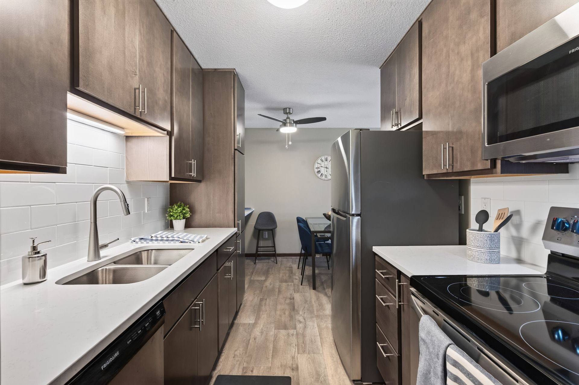 Narrow modern kitchen with dark wood cabinets, stainless appliances, white countertops, and a dining nook beyond.