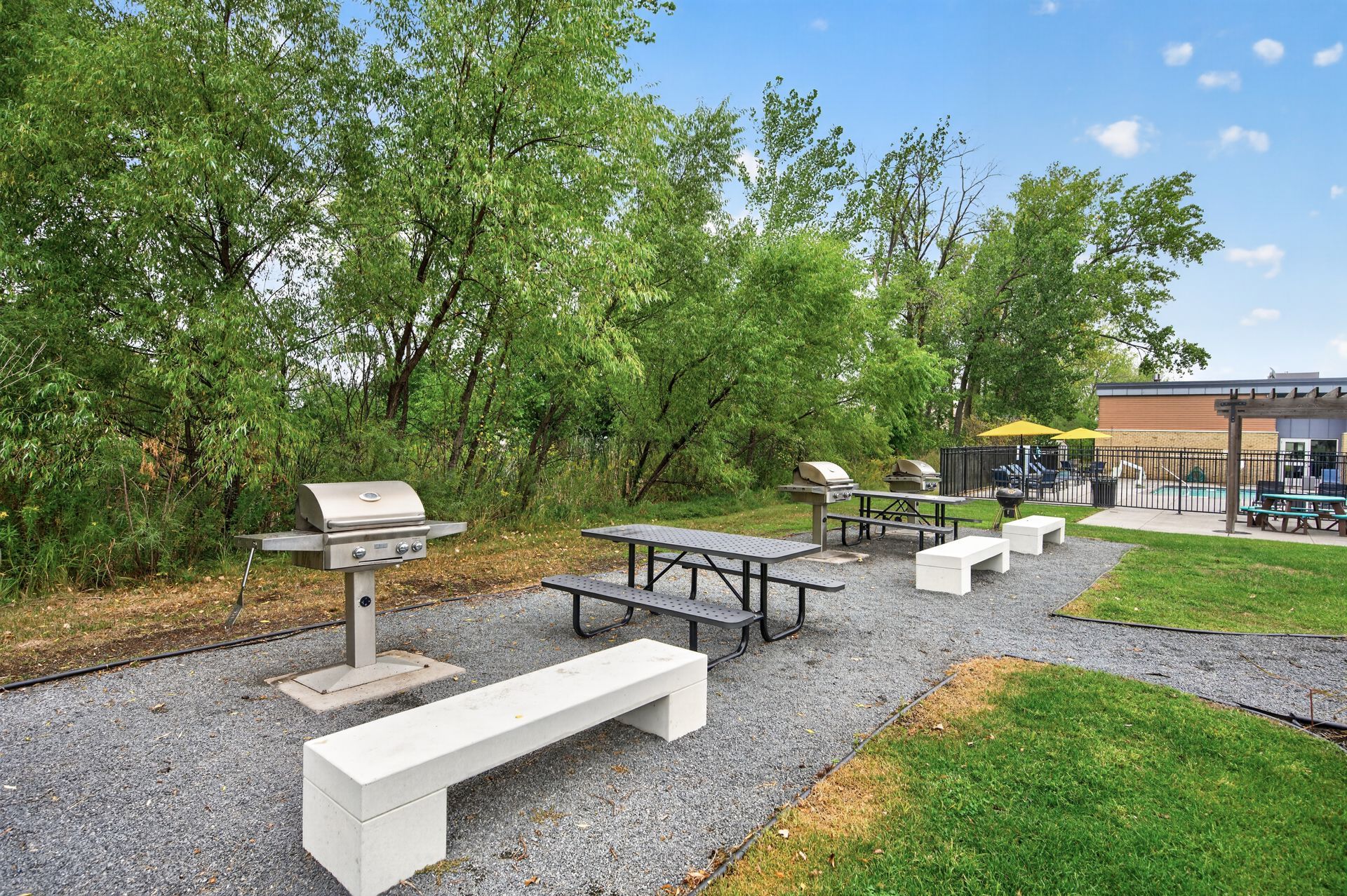 Outdoor picnic area with white benches, tables, a grill, and trees beside a building