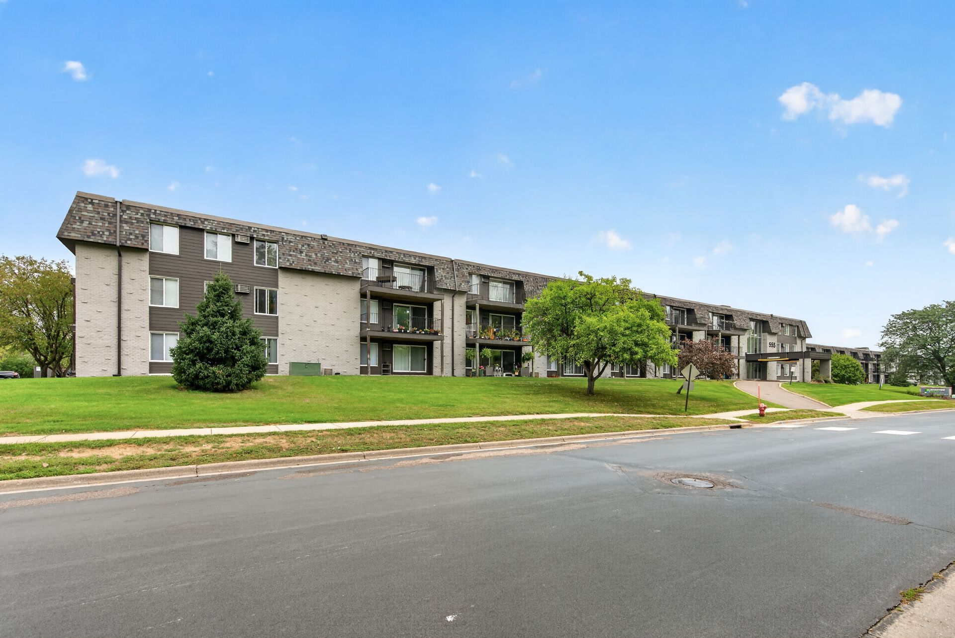 Three-story apartment building with balconies, grassy lawn, and a road in front under a blue sky