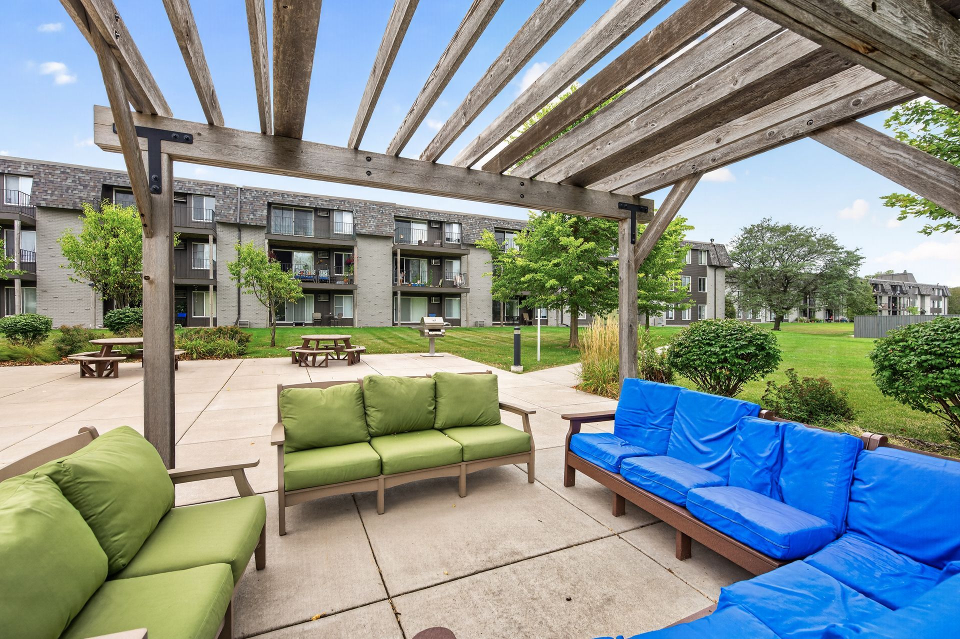 Outdoor patio seating with green and blue cushioned sofas under a wooden pergola, facing an apartment courtyard.
