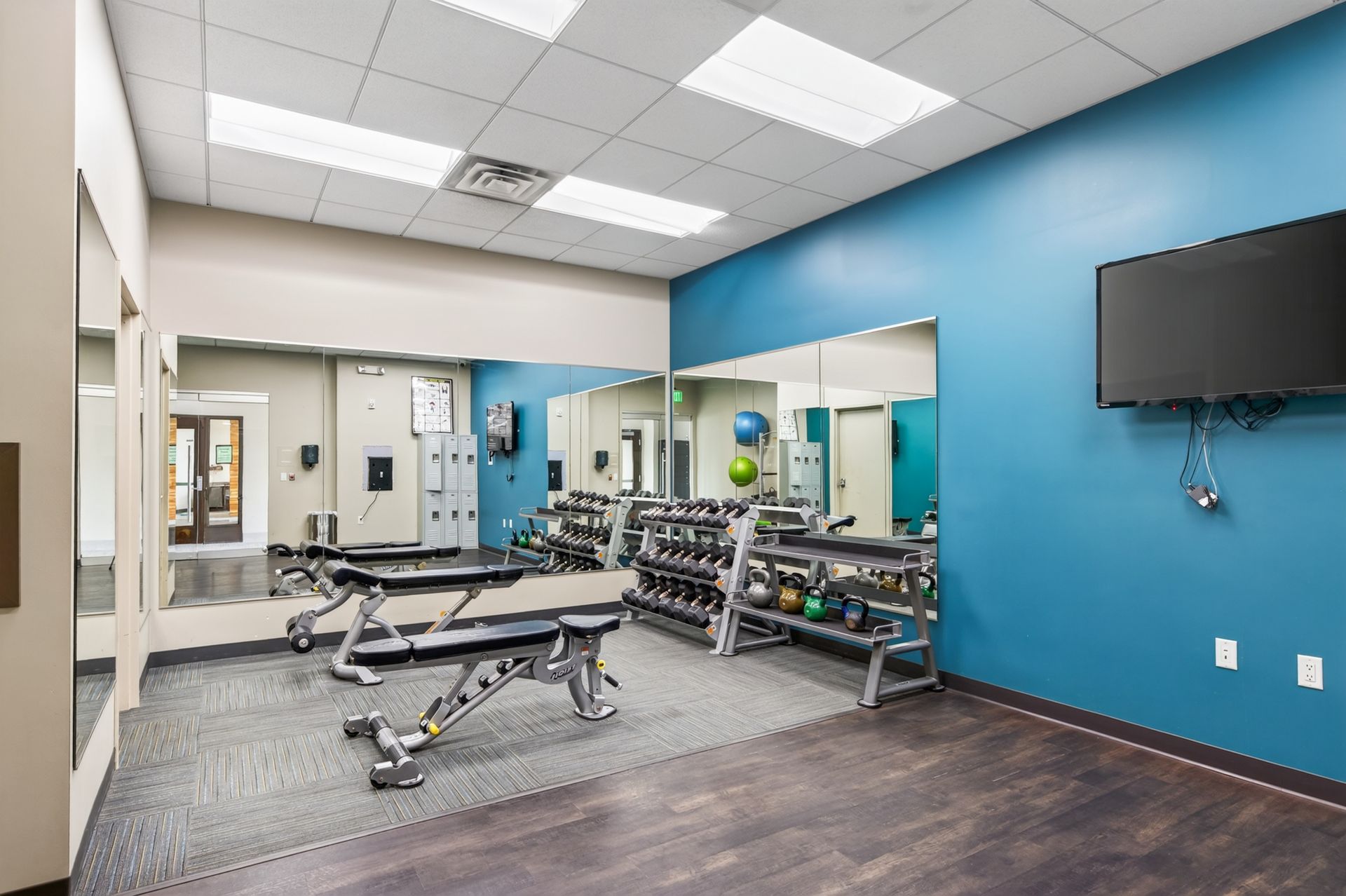 Physical therapy room with exercise benches, dumbbells, mirrors, blue walls, and wall-mounted TV