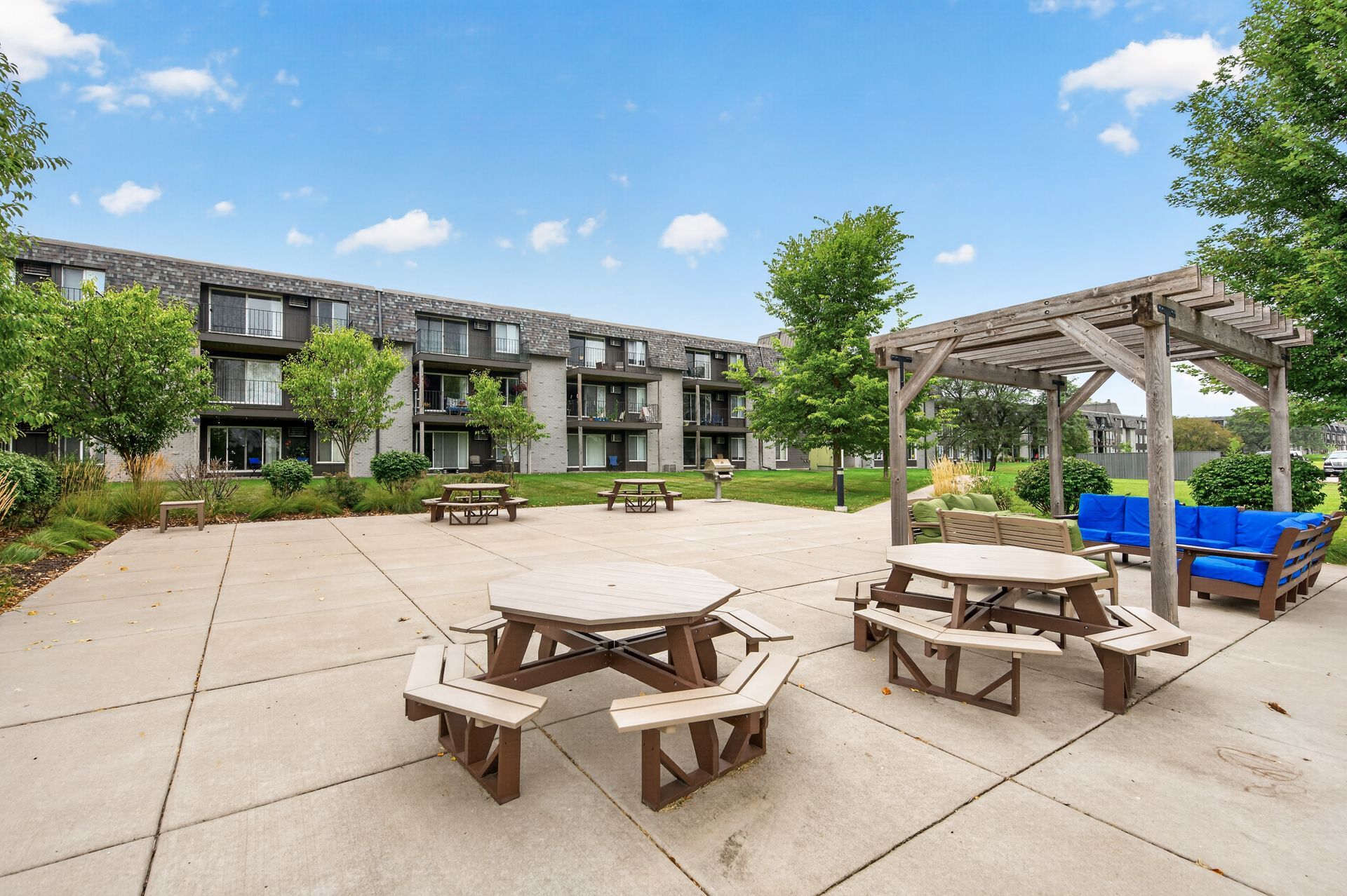 Apartment courtyard with picnic tables, pergola, blue seating, and a building in the background