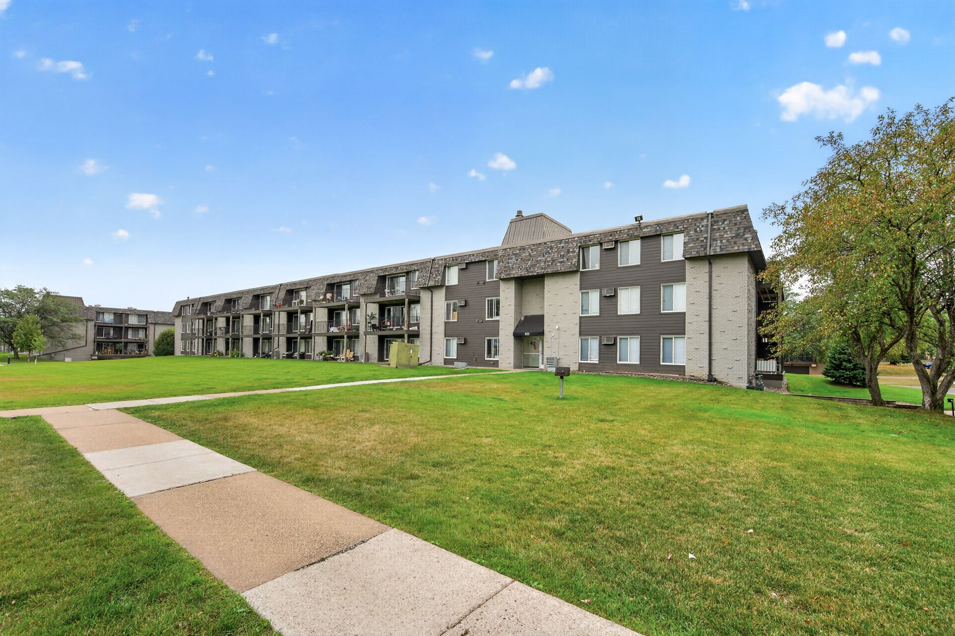 Apartment complex with gray buildings and a large green lawn under a blue sky