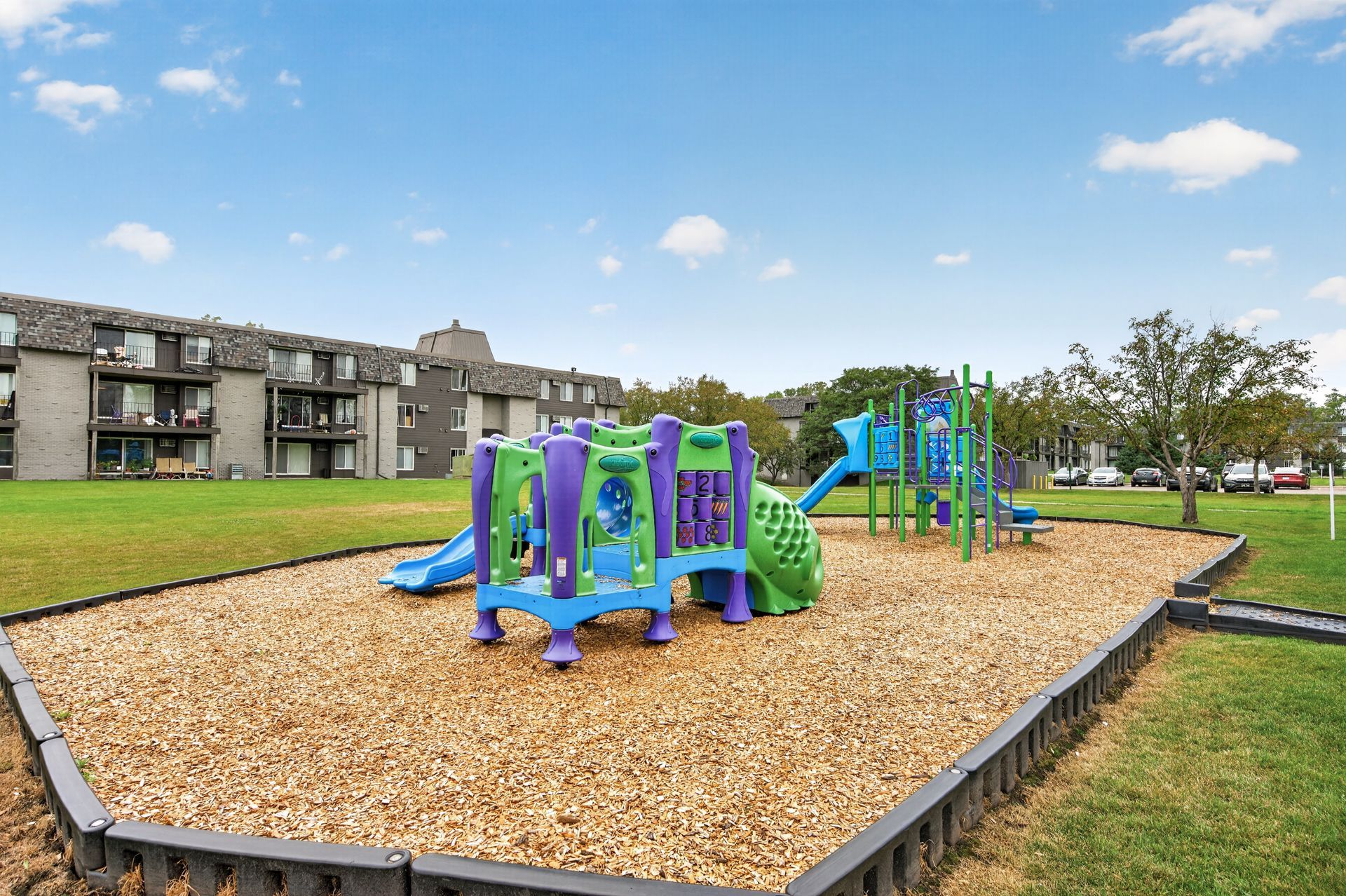 Children’s playground with blue and green climbing equipment on a woodchip play area beside apartments