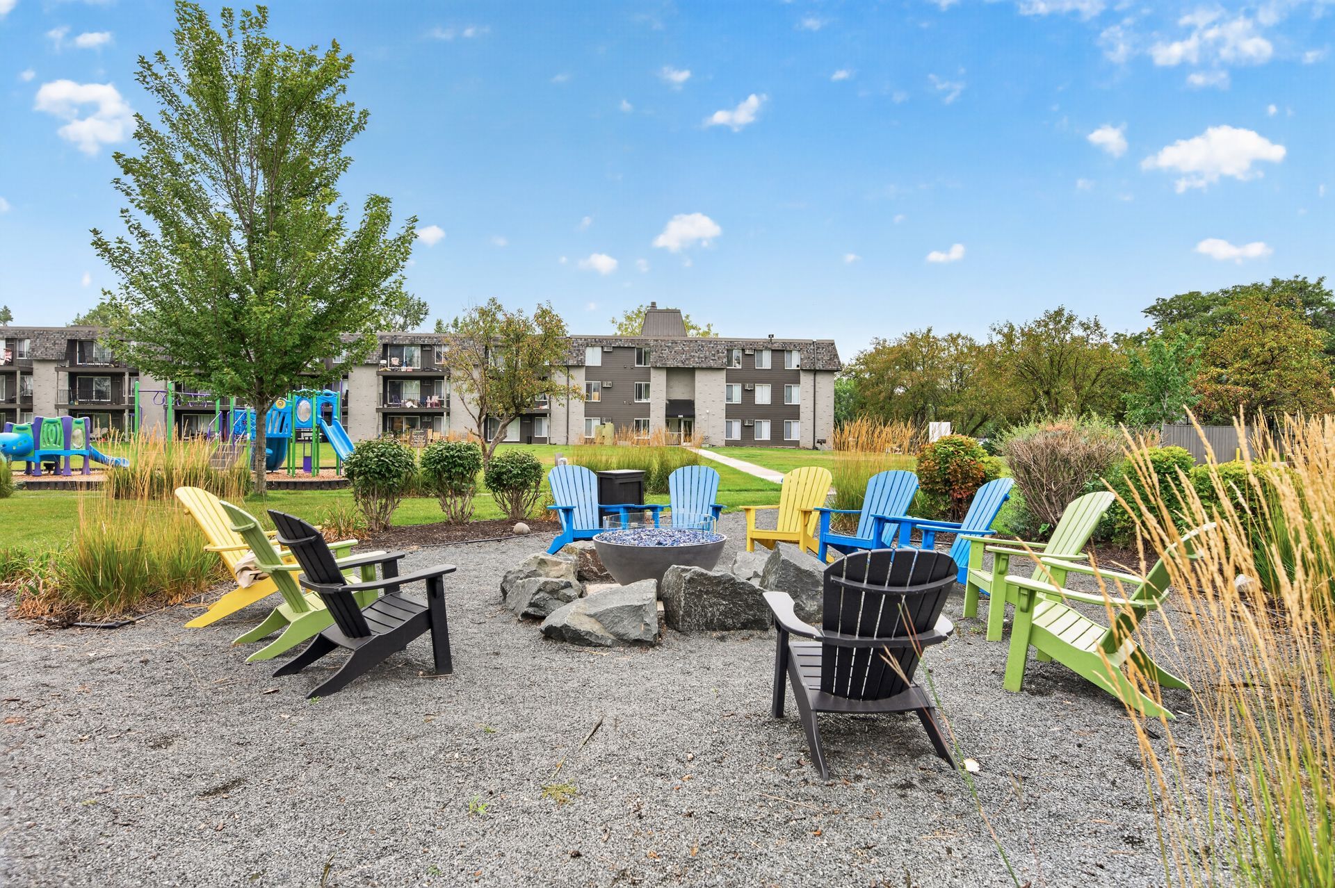 Fire pit seating area with black and green chairs on gravel, surrounded by tall grasses and apartment buildings