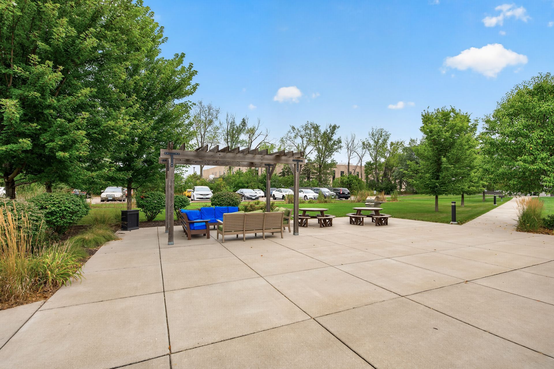 Park walkway with a wooden pergola, blue seating, and surrounding trees on a sunny day