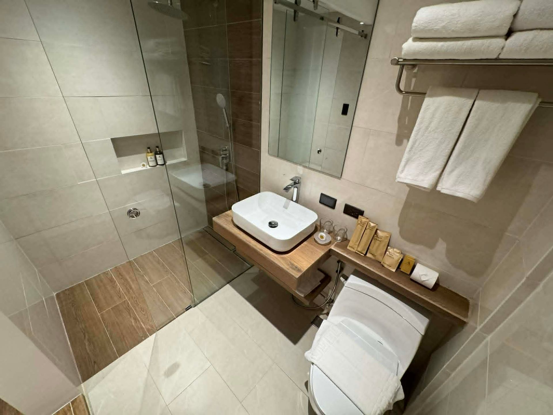 Modern hotel bathroom with glass shower, sink vanity, toilet, beige tile, and folded towels