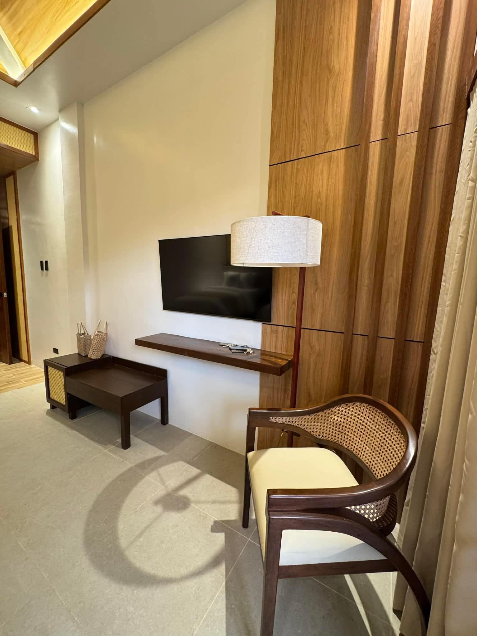 Modern living room with wall-mounted TV, wooden accent wall, table lamp, coffee table, and woven armchair.