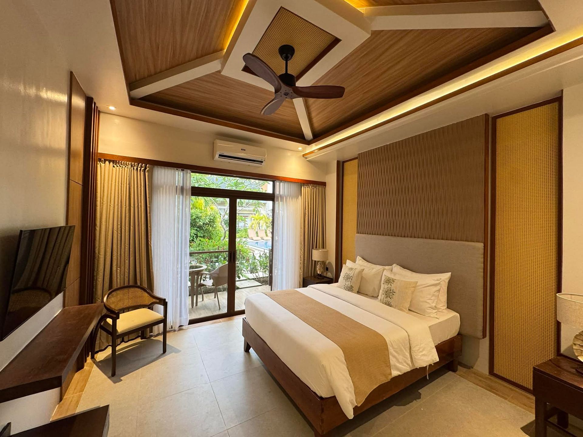 Modern bedroom with a large bed, wood-paneled ceiling, ceiling fan, and balcony doors opening to greenery