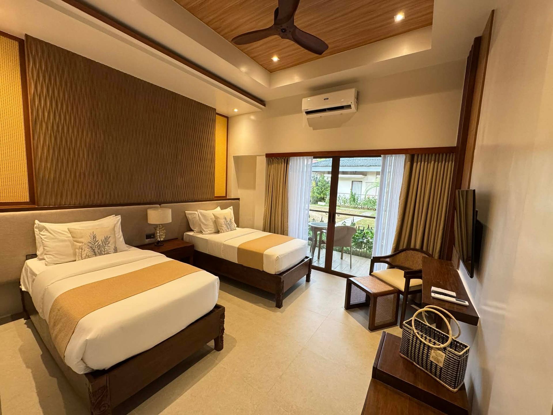 Bright hotel room with two beds, wooden ceiling fan, and balcony doors overlooking greenery