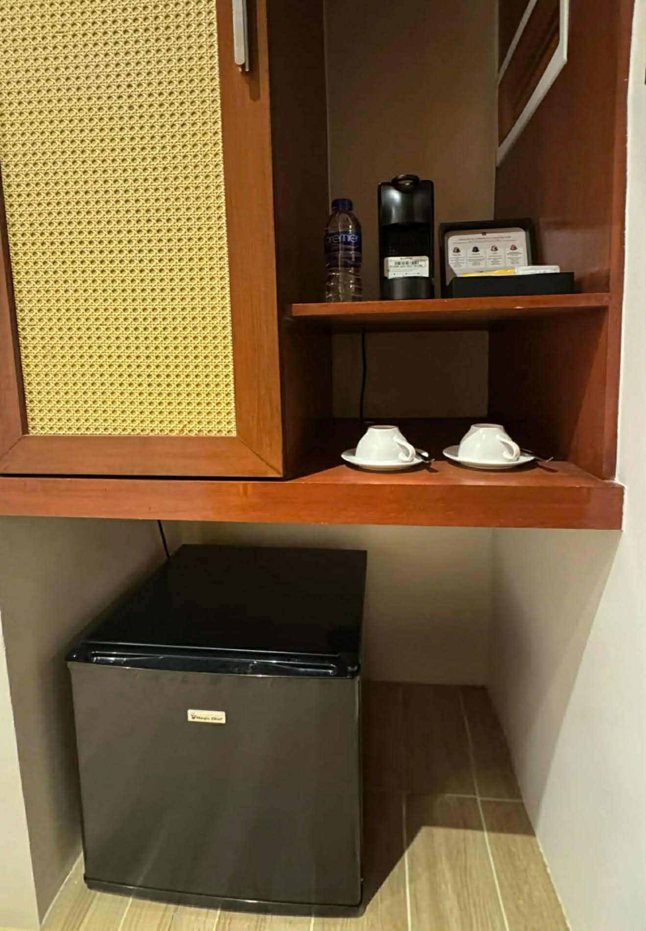 Mini fridge beneath a wooden shelf with cups and bottles in a hotel room nook