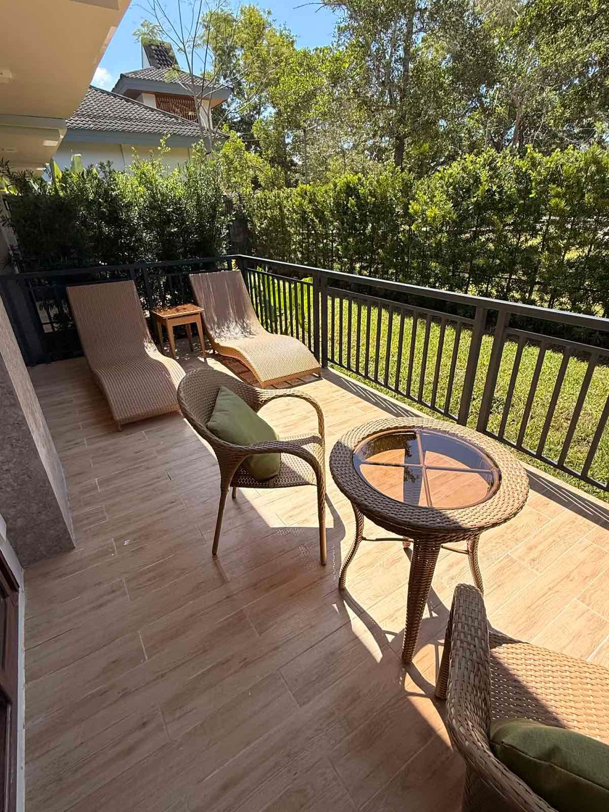 Sunlit wooden patio with lounge chairs, a small table, and trees beyond the railing.