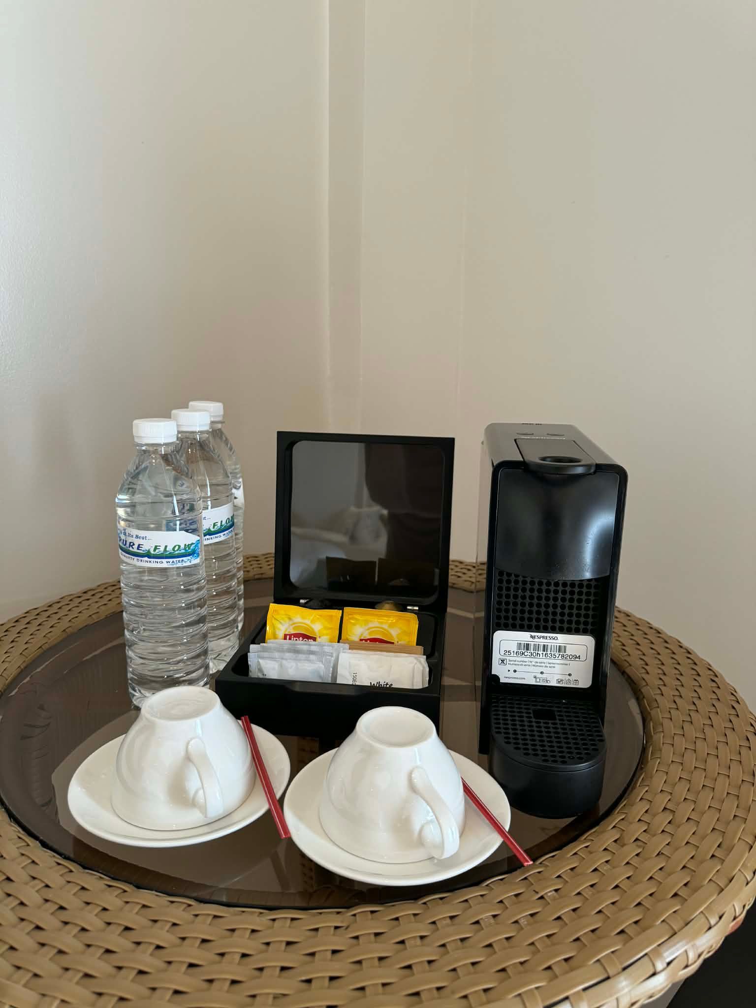 Hotel coffee setup with water bottles, cups, tea sachets, and a black coffee machine on a woven table