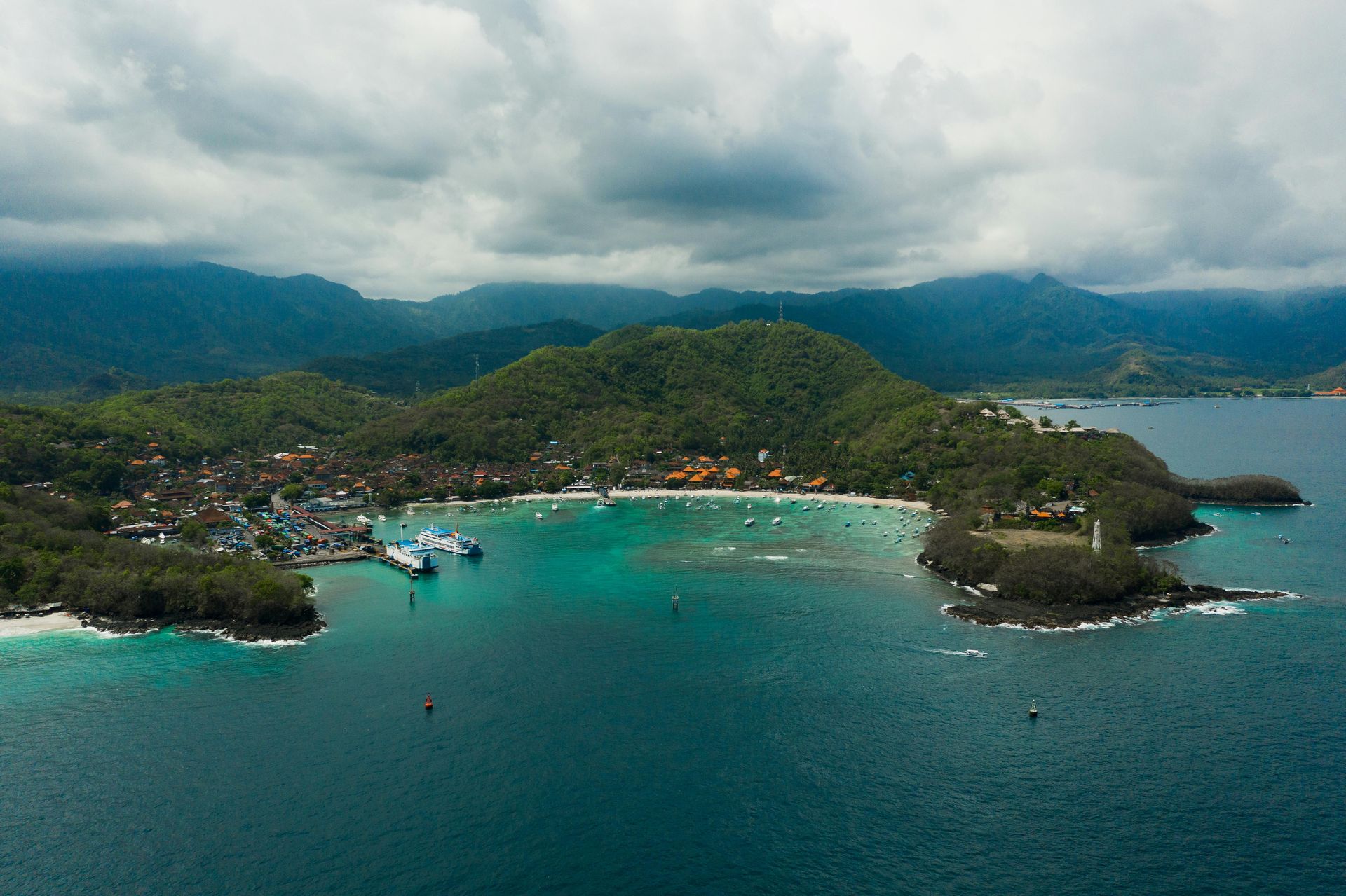 Aerial view of a tropical bay with turquoise water, small boats, and lush green hills under cloudy skies