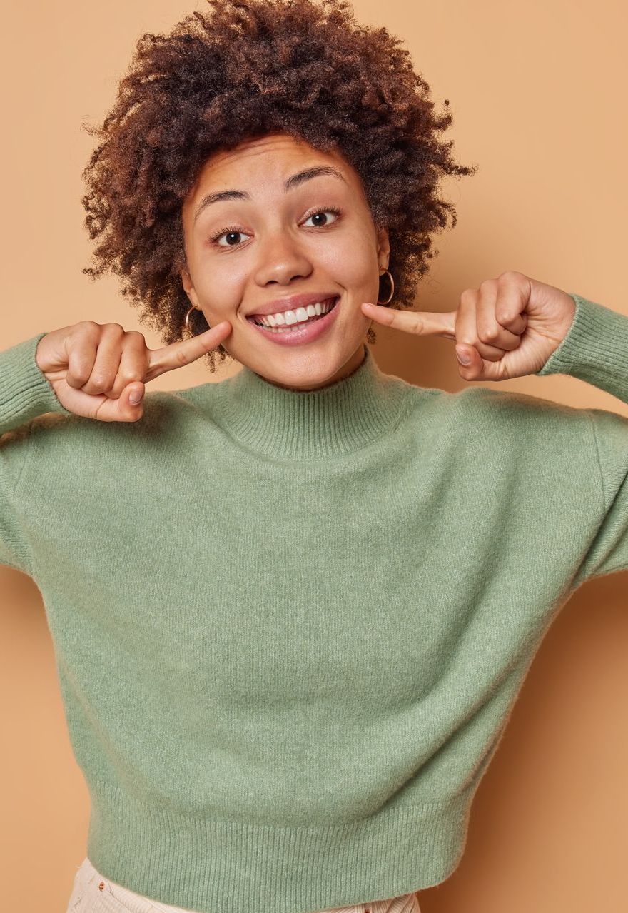 Una donna con un maglione verde sorride e indica i suoi denti. - Arte Dentale Frosinone