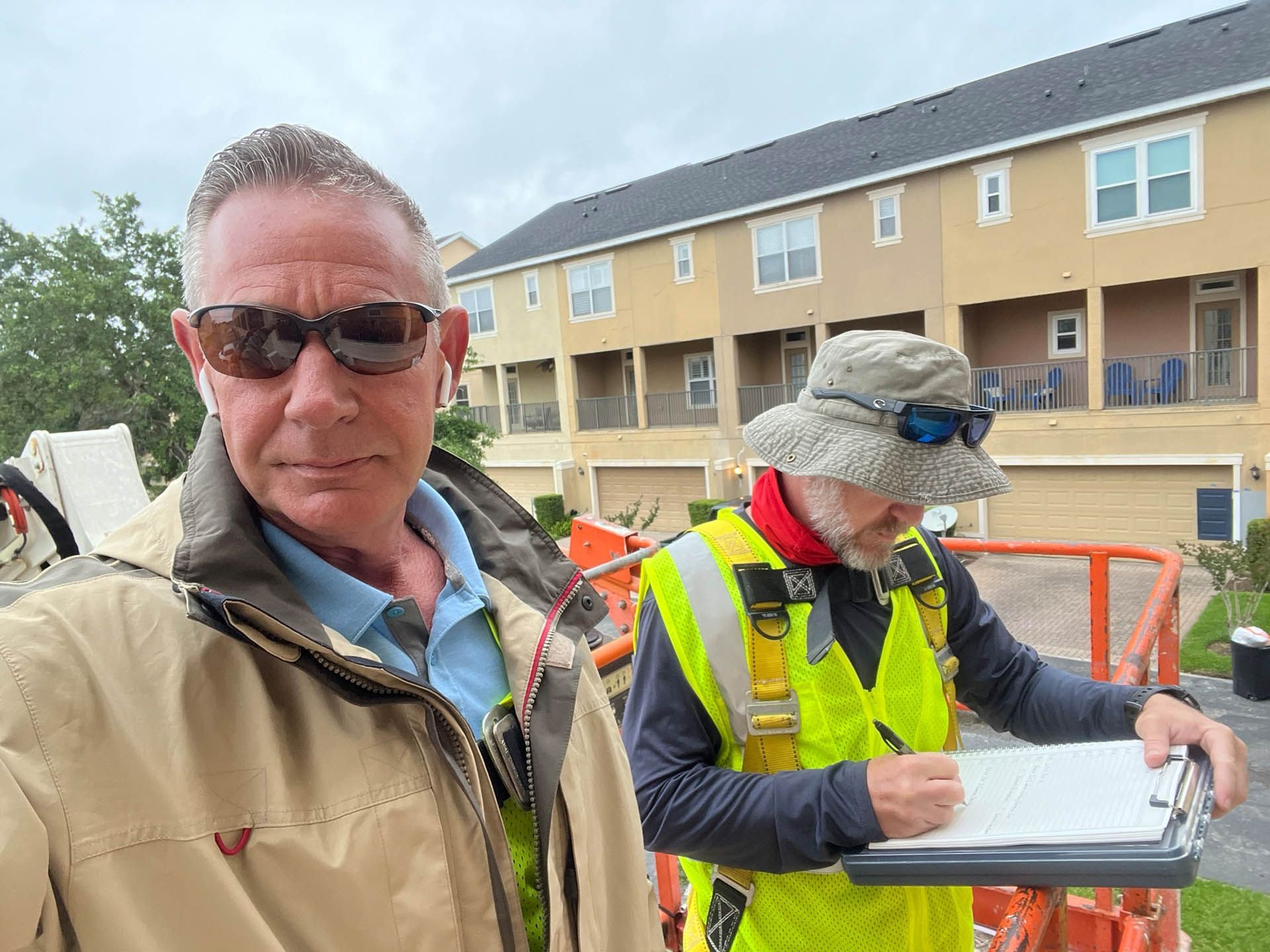 Two people in safety vests on a lift inspect a building exterior. One writes on a clipboard, the other wears sunglasses.