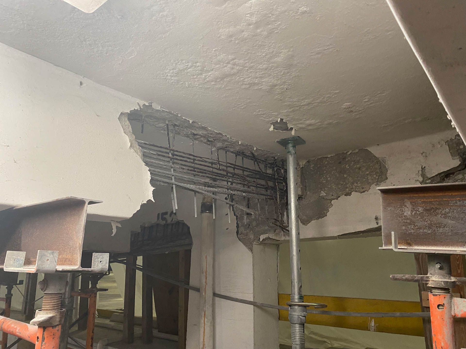Damaged concrete ceiling with exposed rebar, supported by metal beams and props in a construction setting.