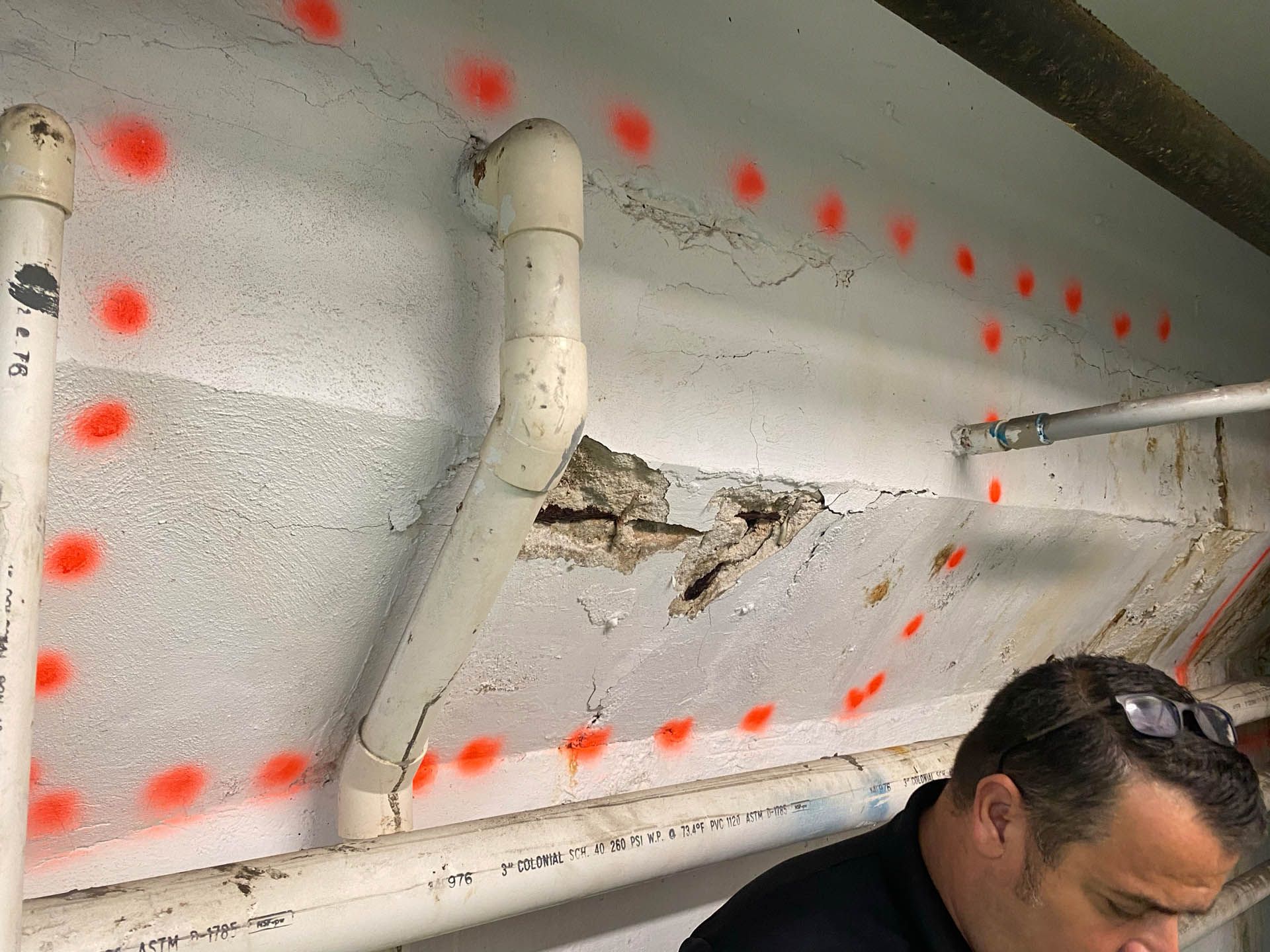 Damaged concrete ceiling with exposed rebar, orange spray-painted markings, and a person observing.