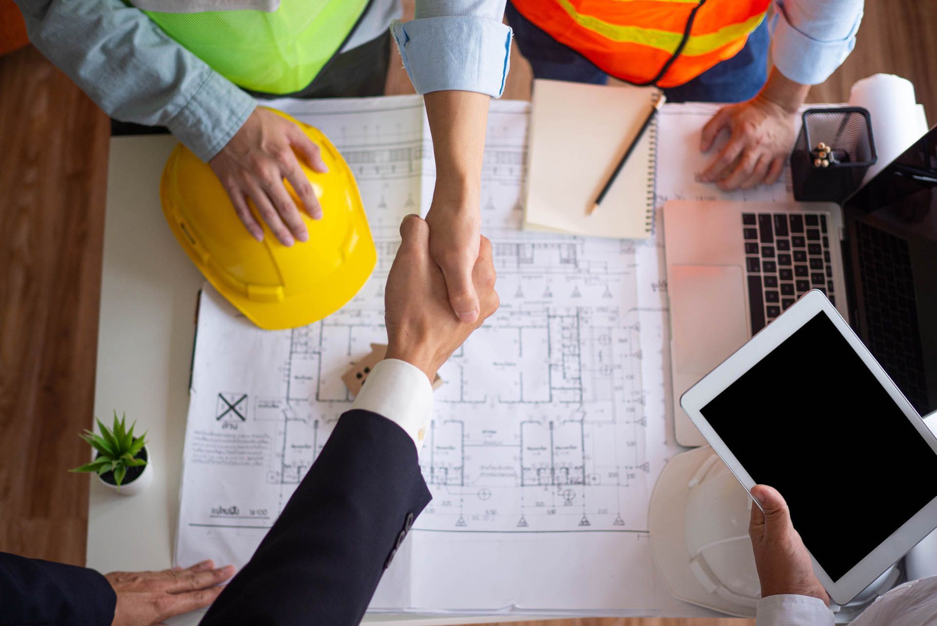 Two people shaking hands over blueprints, a laptop, and a tablet, with hard hats and safety vests visible.