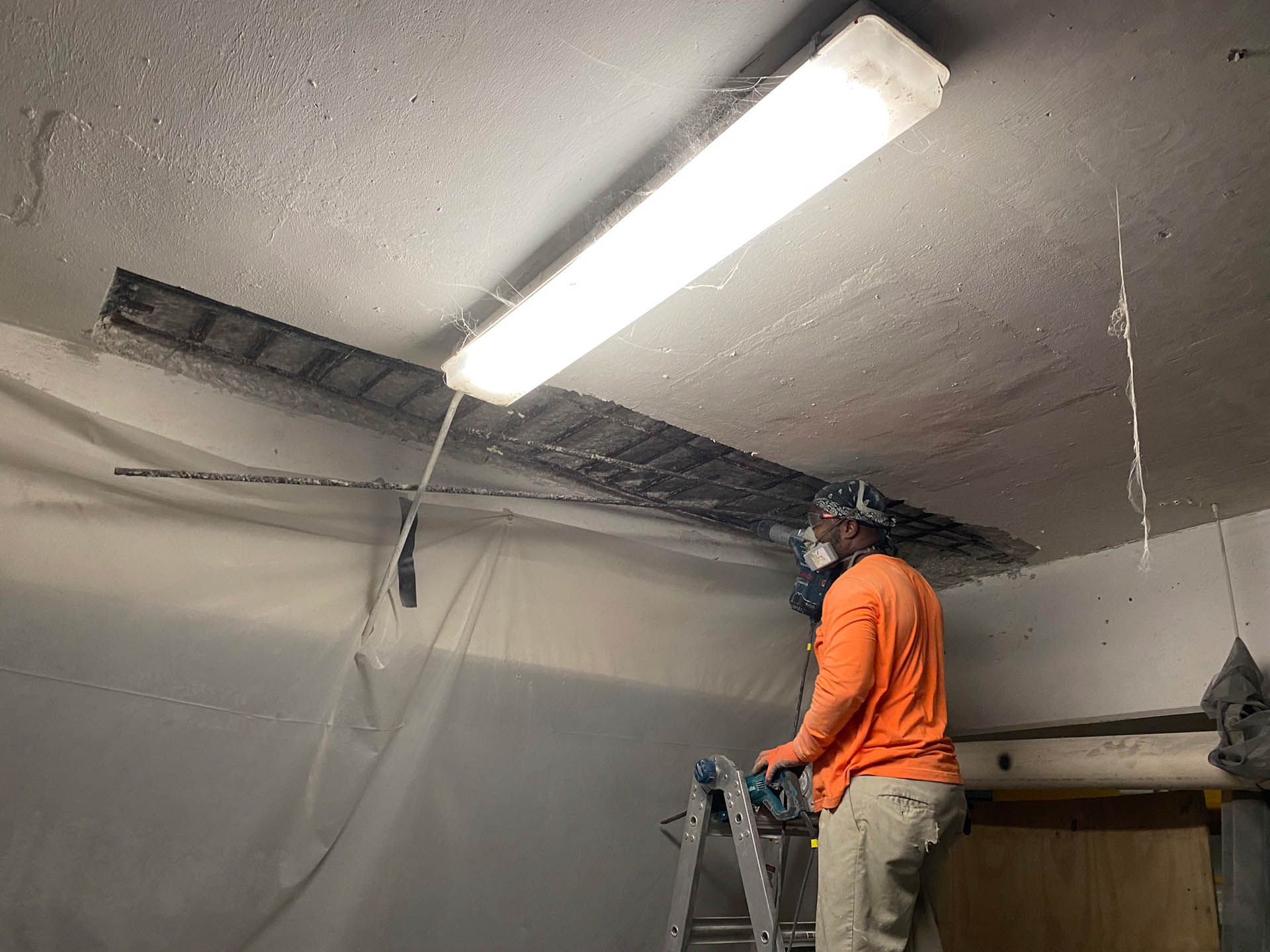 Person in orange shirt and respirator on ladder, working on ceiling with overhead light.