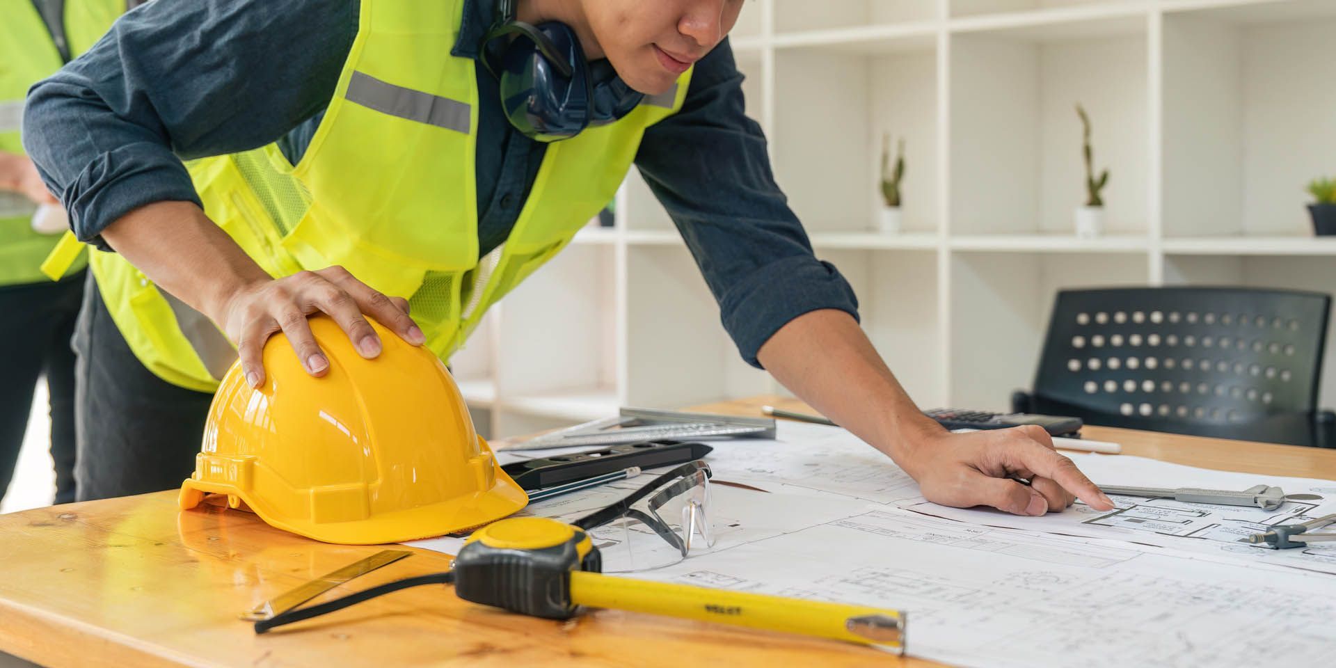 Person in safety vest and hard hat examines blueprints at a desk with tools and office shelves in the background.
